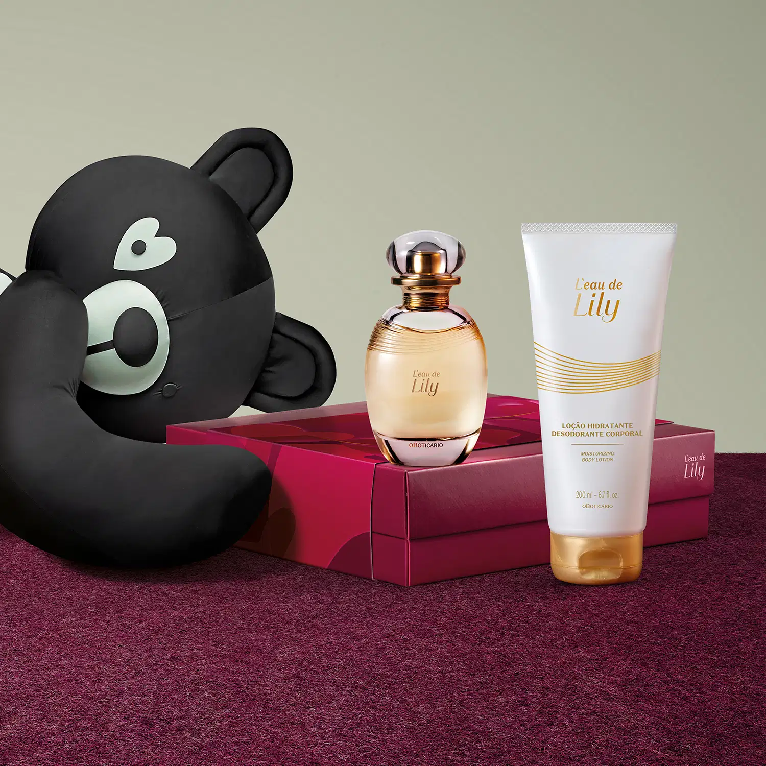 Urso de tecido preto e branco, frasco de desodorante colônia Lily, frasco de loção hidratante corporal da mesma marca e caixa na cor vinho sobre superfície vermelha, em ambiente minimalista com tons neutros.