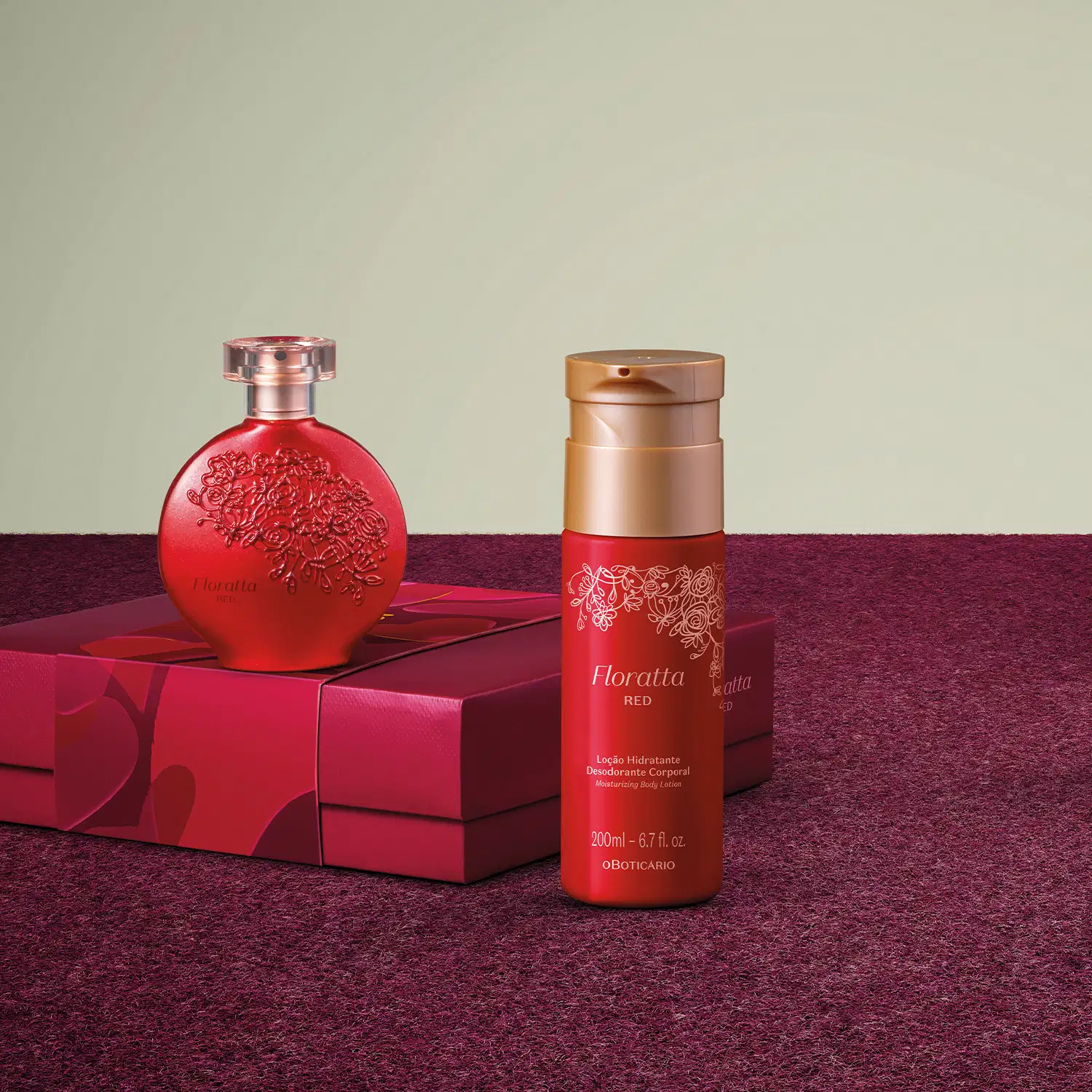 Frasco vermelho e dourado de loção hidratante corporal, frasco de 
 desodorante colônia em embalagem vermelha arredondada e caixa vinho sobre superfície vermelha com detalhes e fundo cinza.