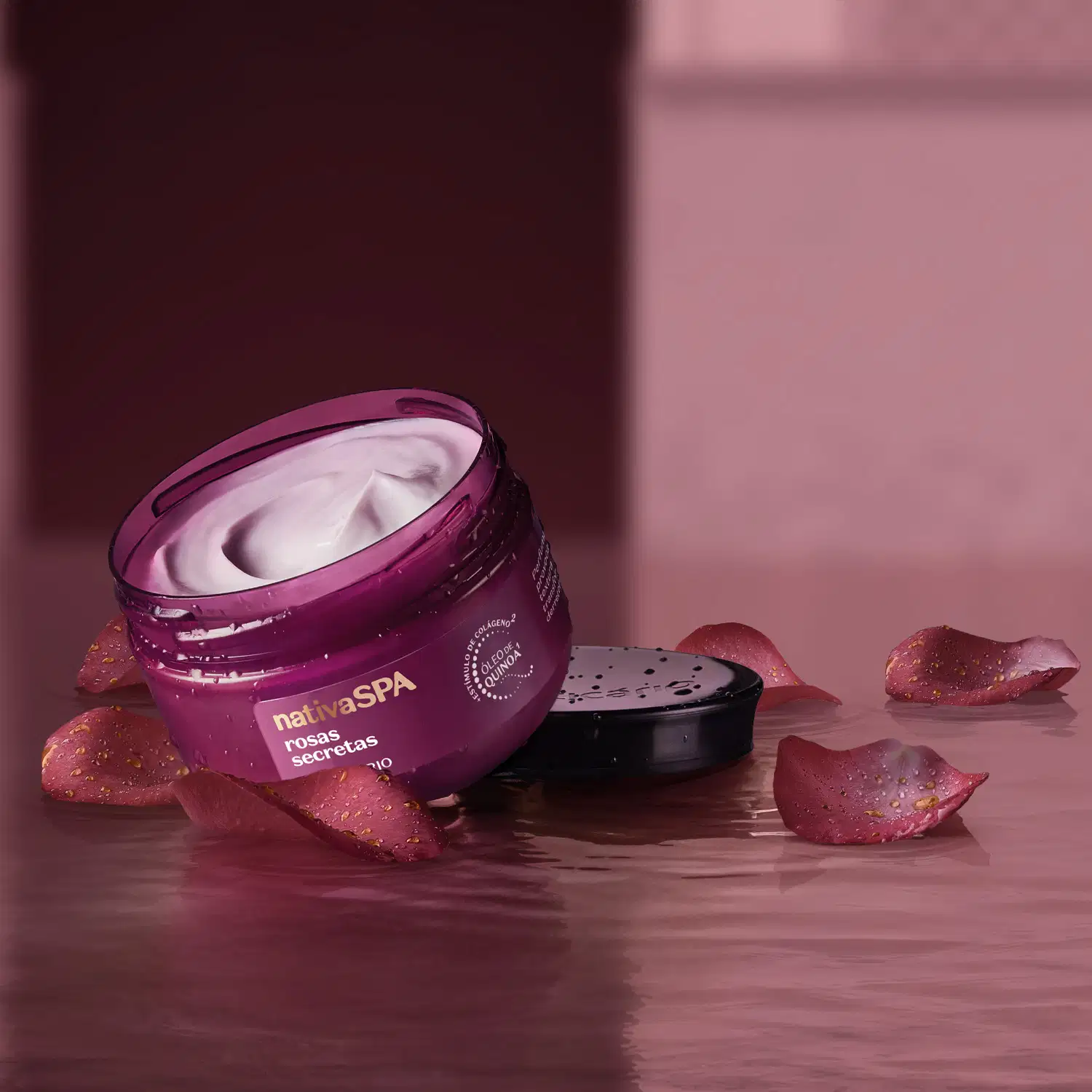 Ambiente de spa com um pote de óleo em creme Nativa SPA Rosas Secretas. Ele está sobre pétalas de rosa espalhadas sobre uma superfície úmida, em um cenário monocromático de tons de roxo.