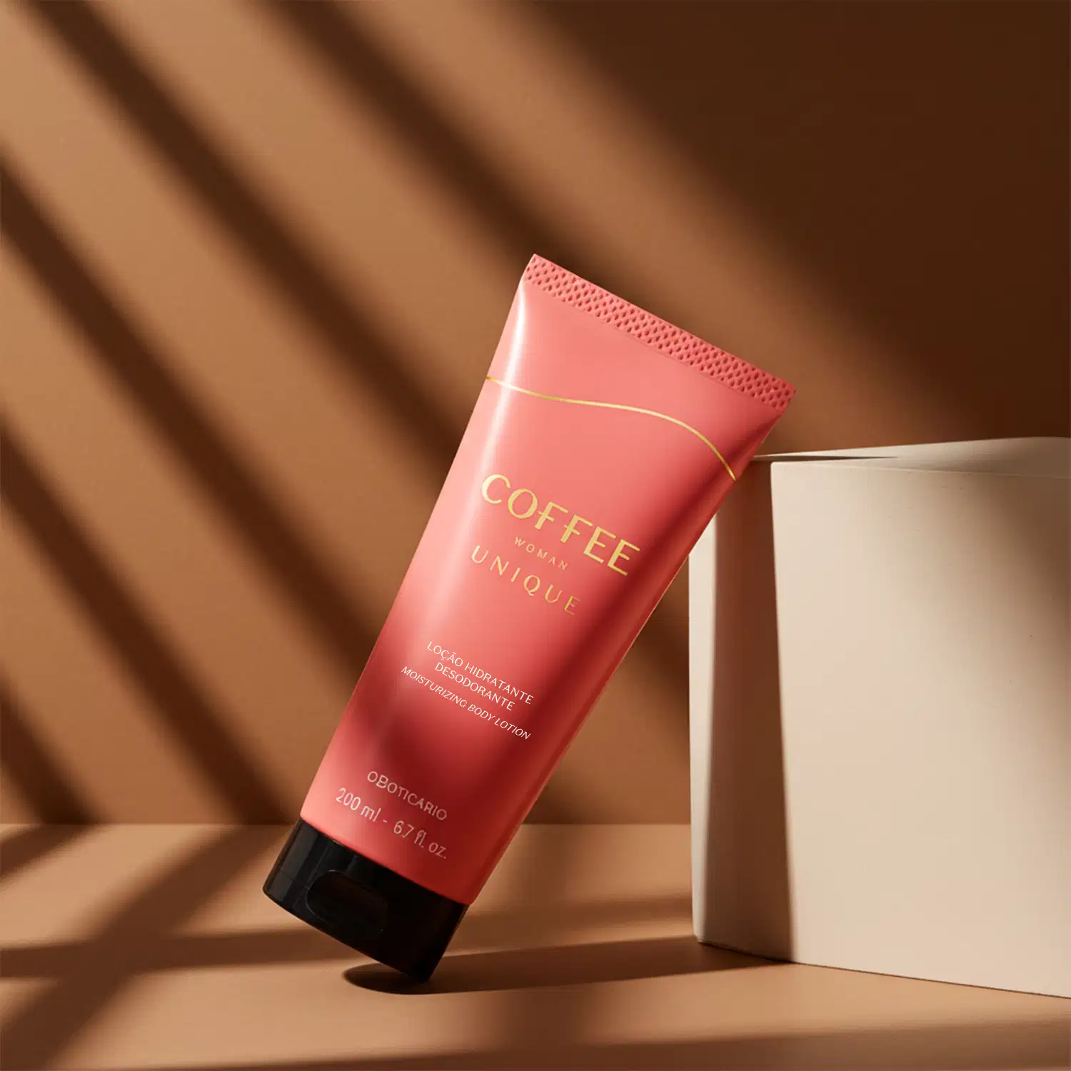 Imagem de uma bisnaga de loção hidratante desodorante da linha Coffee Woman Unique com embalagem rosa e tampa preta reclinada sobre um cubo rosa claro e apoiada em superfície e fundo bege com sombras diagonais projetadas por iluminação natural.