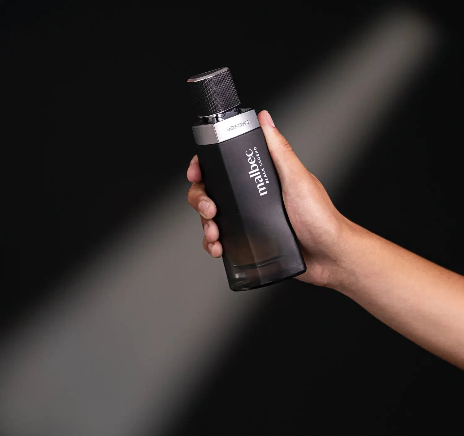 Imagem de um frasco preto de desodorante colônia Malbec Black Legend em uma mão contra um fundo escuro. O produto possui um design minimalista e elegante.