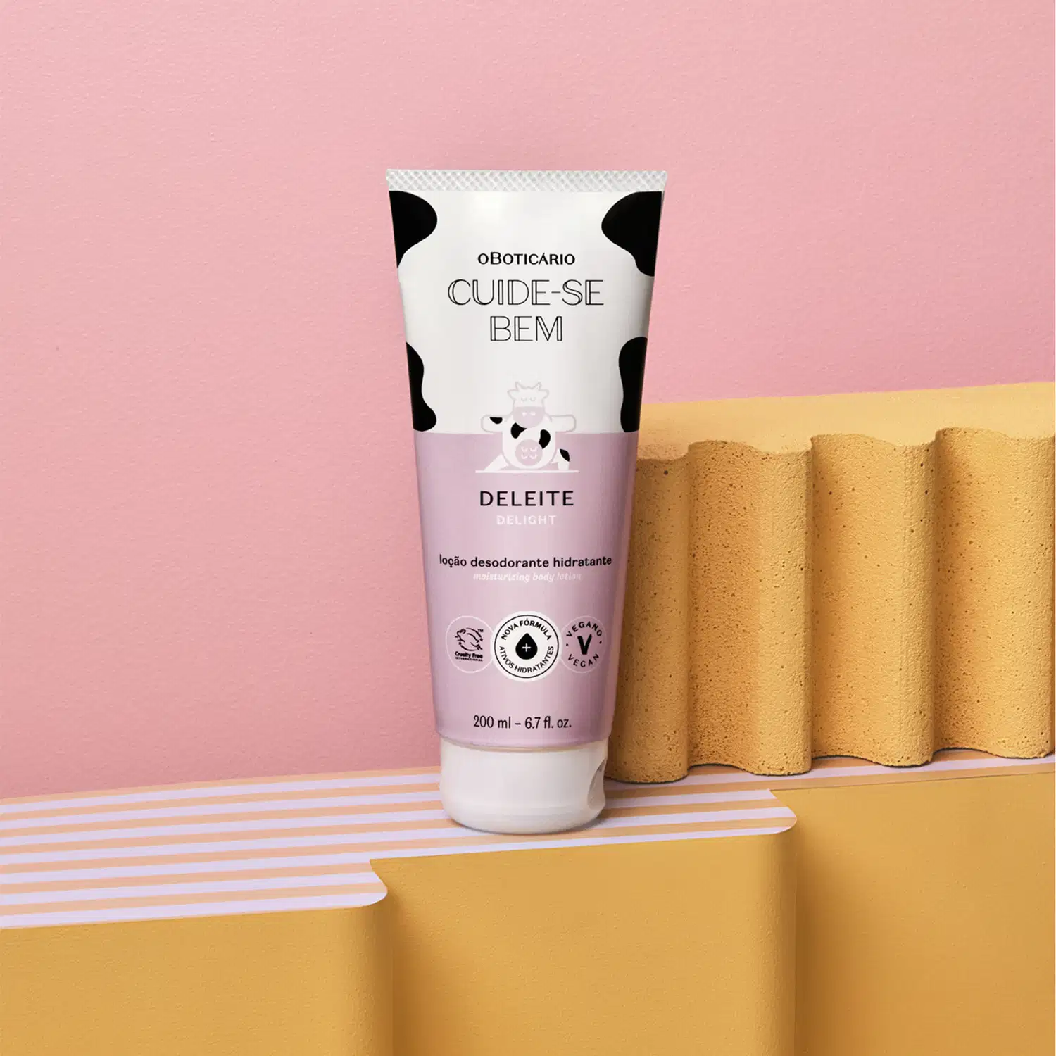 Tubo branco de desodorante hidratante da linha Cuide-se Bem Deleite, da marca Oboticario posicionado sobre superfície ondulada amarela com detalhes listrados de rosa e branco e fundo rosa.