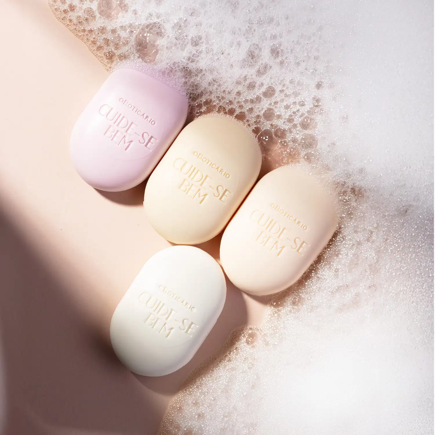 Imagem de quatro sabonetes sortidos da marca Cuide-se Bem em tons de lilás e rosa sobre superfície rosa com espuma branca, sugerindo um ambiente de cuidado pessoal e bem-estar.