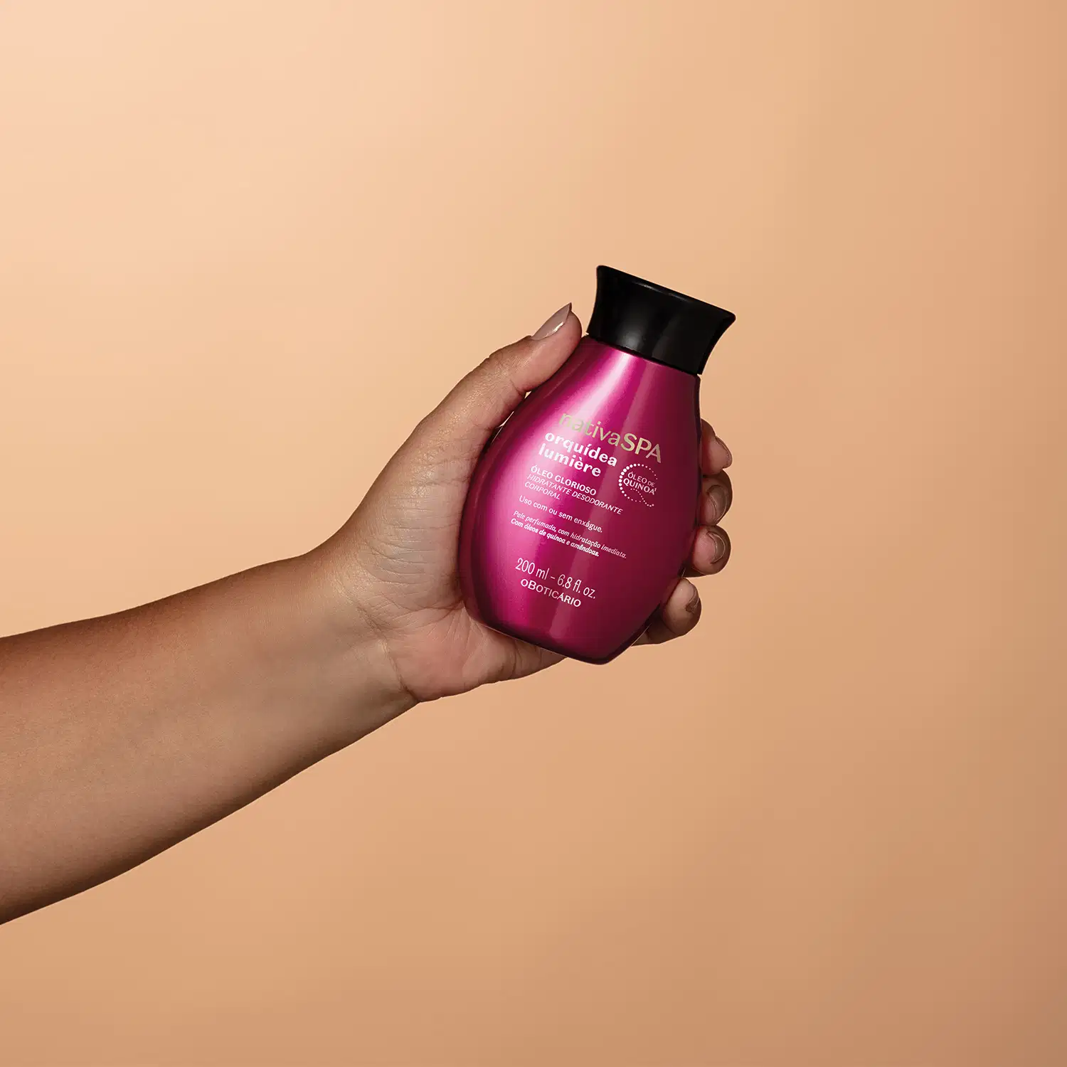 Imagem mostra mão e antebraço femininos segurando uma embalagem pink Oleo Glorioso Nativa SPA Orquídea Lumiere em fundo laranja claro.