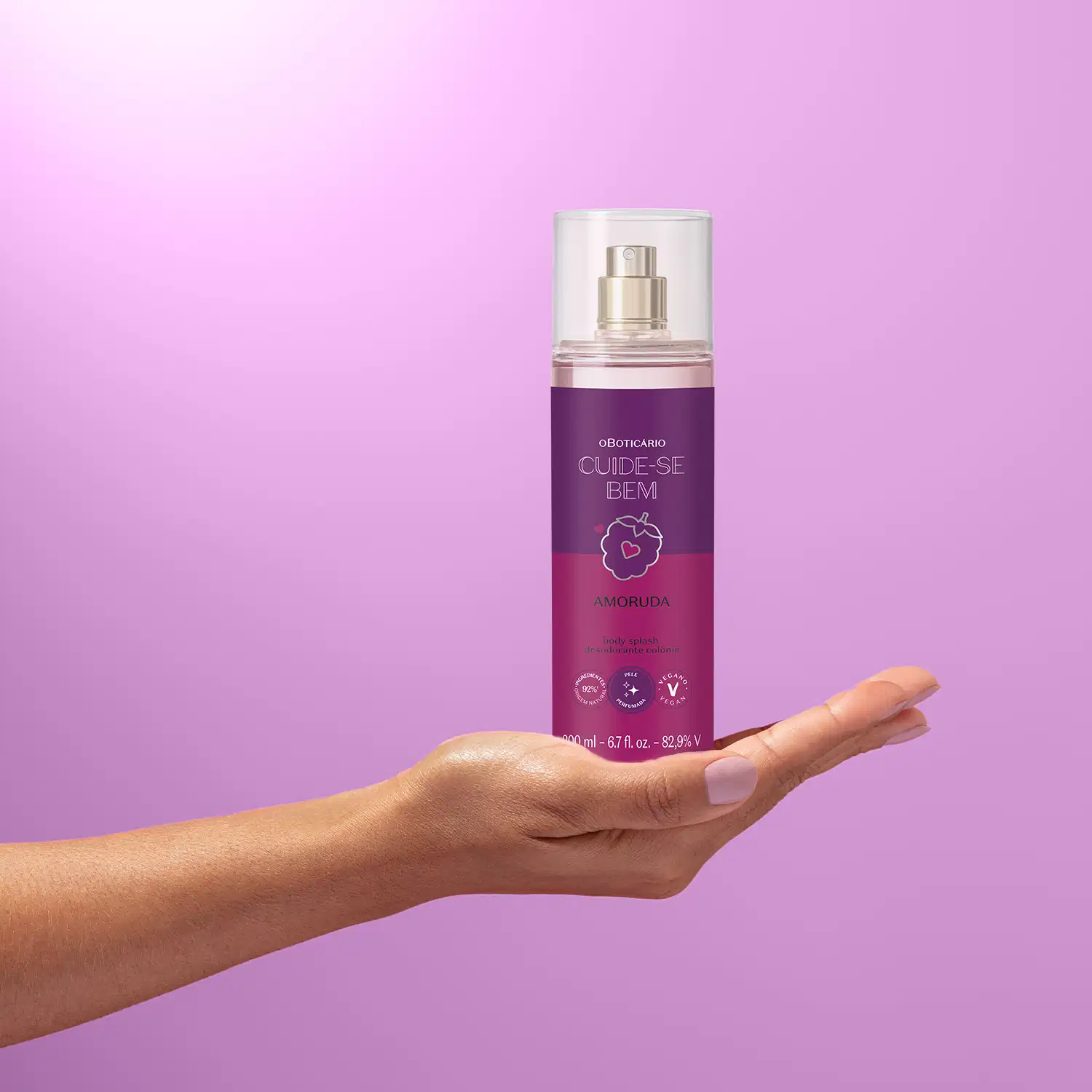 Mão segurando um frasco de body splash em embalagem transparente com líquido lilás e rótulo roxo da marca Cuide-se Bem Amoruda em fundo lilás.