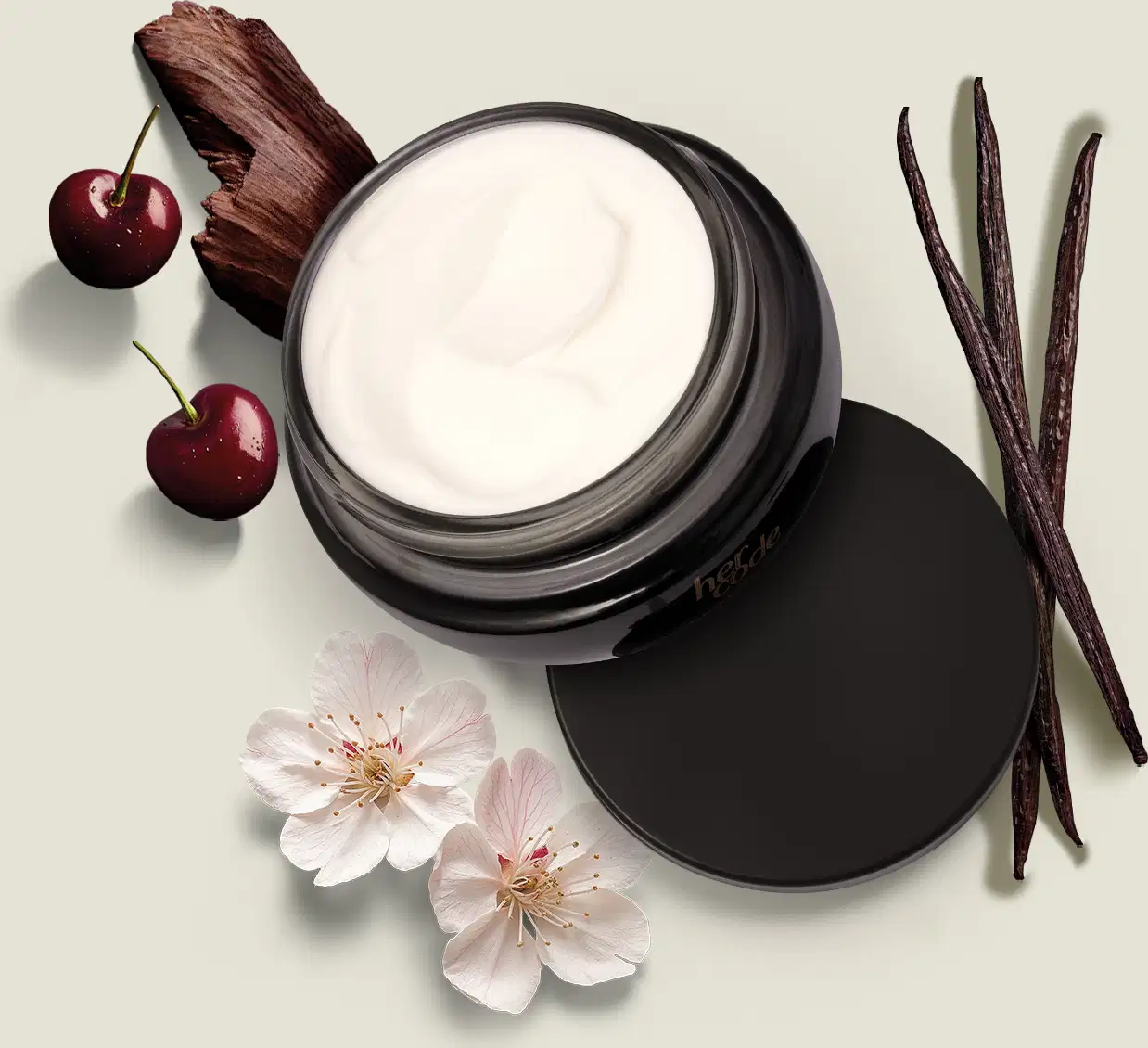 Ao redor do pote preto com creme branco contendo creme hidratante corporal da marca Her Code, há alguns elementos decorativos, como cereja, flores e uma vagem de baunilha. A imagem tem um fundo claro e neutro, destacando o produto central.
