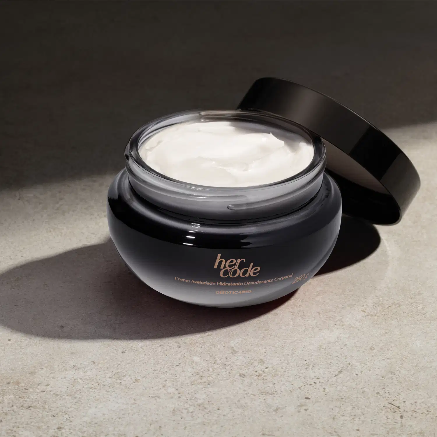 O produto é um creme hidratante desodorante da linha Her Code, em uma embalagem de vidro com tampa preta. O creme em si tem uma aparência cremosa e branca, preenchendo o recipiente de vidro. Está em fundo escuro com iluminação na superficie.