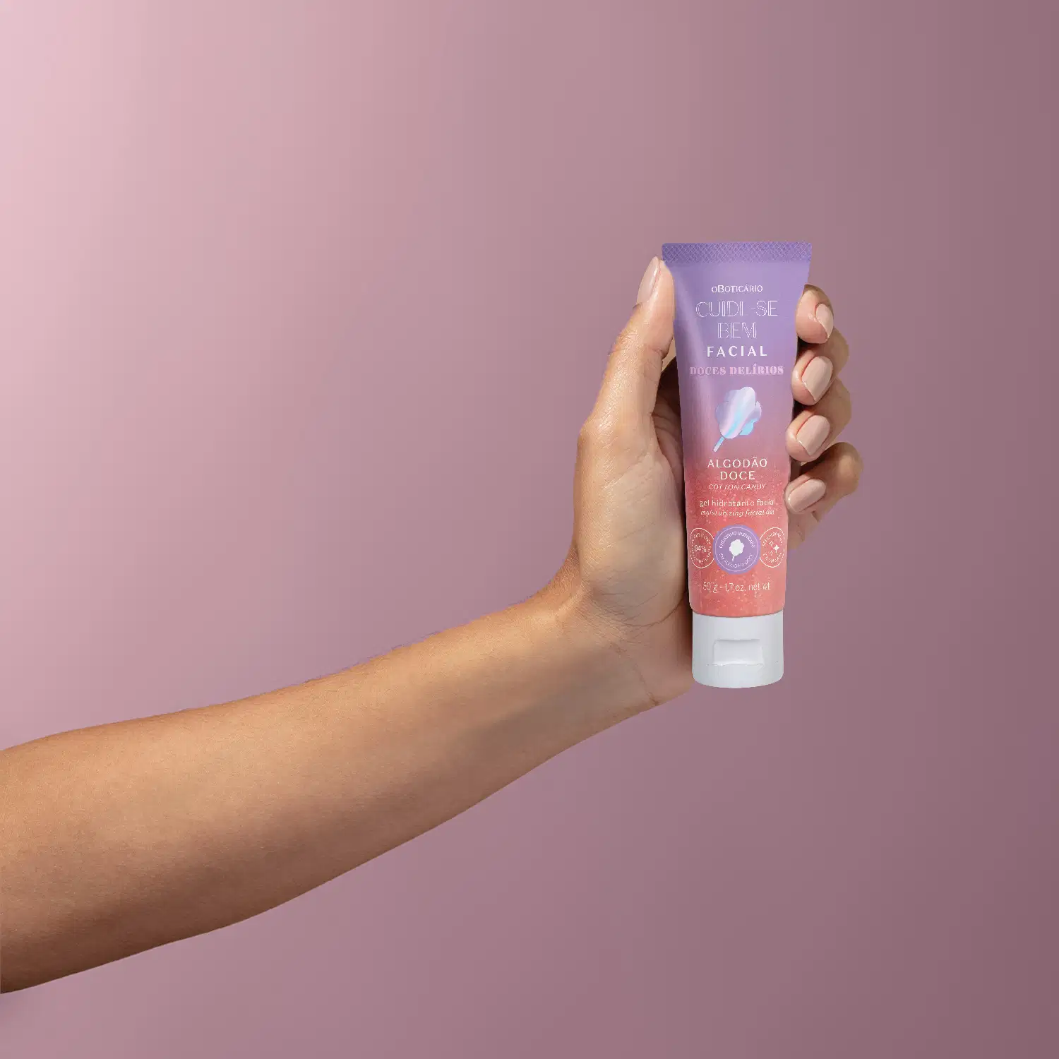 Imagem de uma mão segurando um frasco de gel hidratante facial da marca Cuide-se Bem Algodão Doce, com embalagem em tons de roxo, rosa e branco em fundo com degradê do lilás para o roxo.