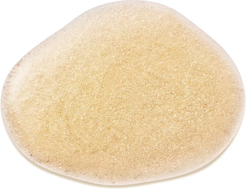 Objeto de textura granulada, com coloração amarelo-clara. Aparenta ser um produto de beleza, possivelmente um item de perfumaria. Sua superfície apresenta uma consistência sólida e brilhante, sugerindo uma textura arenosa ou cristalina.