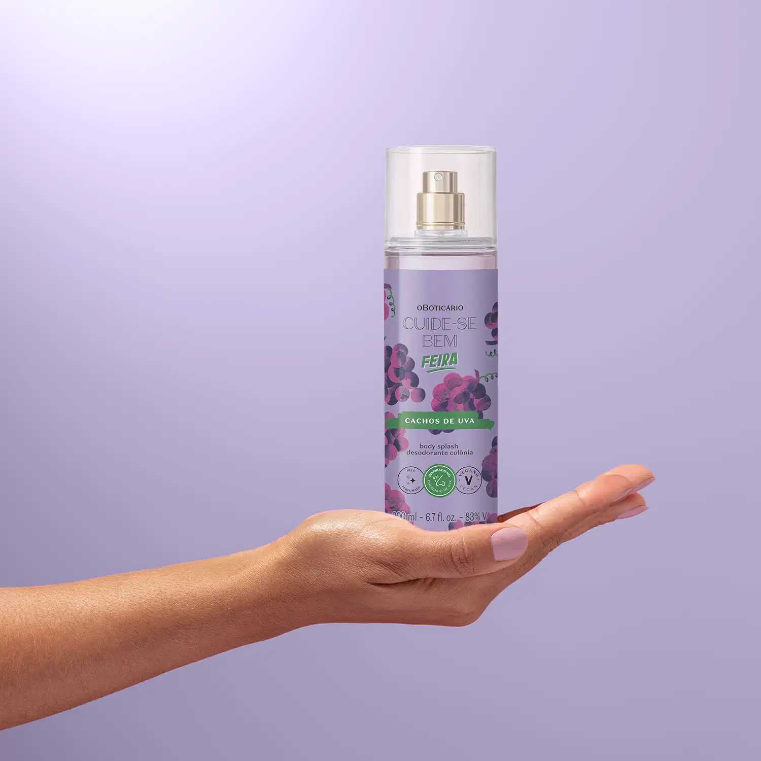Imagem de um frasco de body splash da marca Cuide-se bem Feira Cachos de Uva com rótulo roxo e ilustração de uvas roxas, seguro por mão em fundo roxo claro.