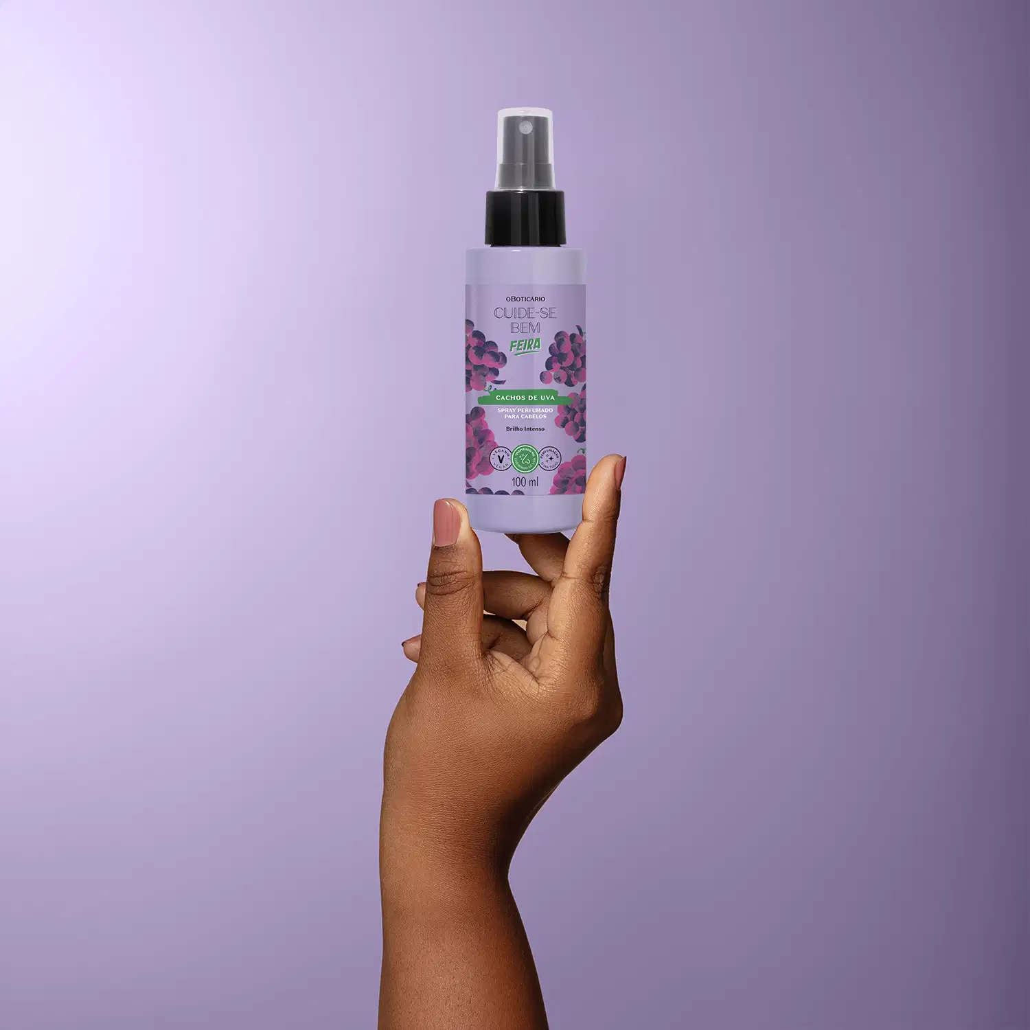 Frasco roxo de spray perfumado para cabelos com rótulo roxo e imagem de uvas, em mão contra fundo roxo claro.