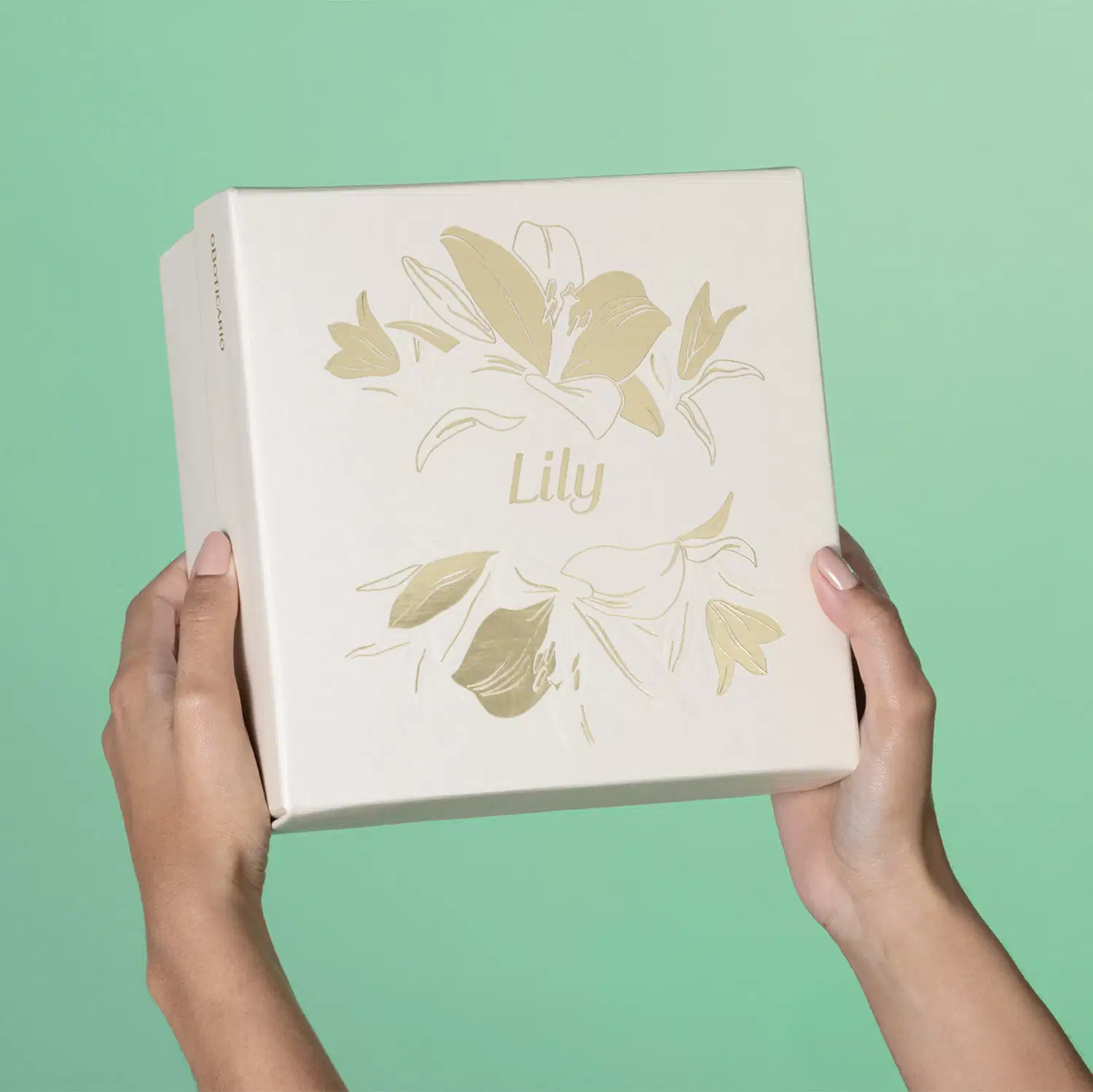 Imagem de uma caixa branca com ilustração dourada de flores, sobre fundo verde claro, segura por mãos femininas.