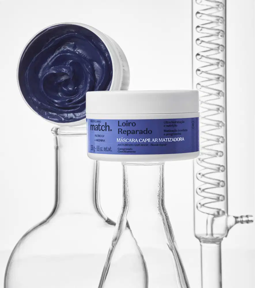 Imagem de um pote branco com rótulo azul da marca Match contendo máscara capilar reparadora, sobre superfície clara com frascos de vidro laboratoriais ao redor. Também mostra a embalagem aberta apresentando um creme azul escuro.