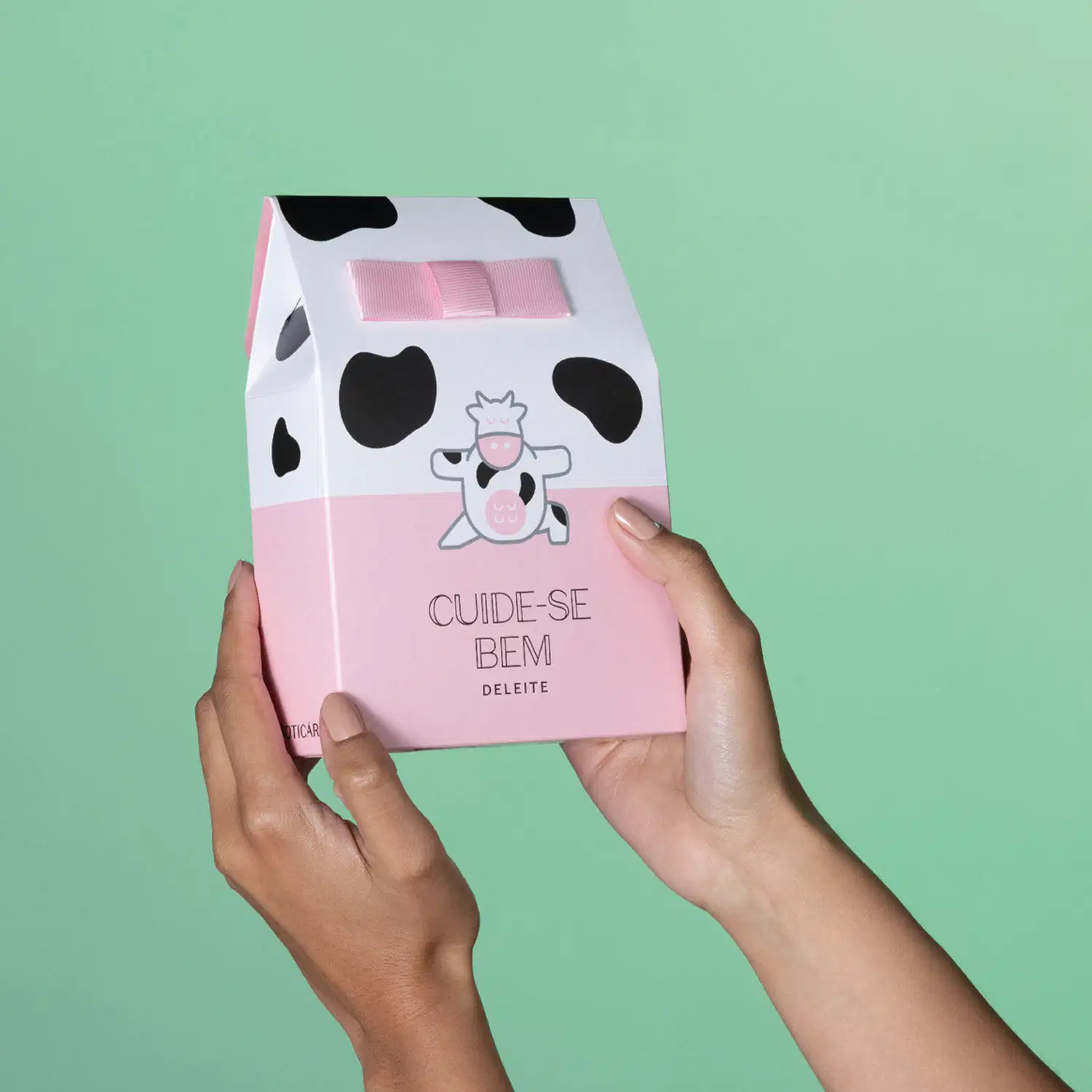 Imagem de uma mão segurando uma sacola presenteável branca e rosa com manchas pretas e ilustração de vaquinha da marca Cuide-se Bem Deleite em fundo verde.