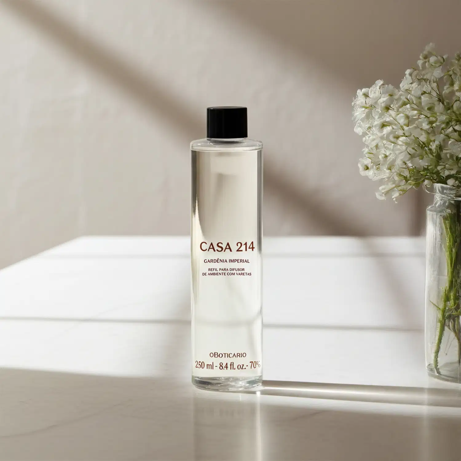 Imagem de um frasco refil com a fragrância Gardênia Imperial para difusor de ambiente da marca Casa 214 em embalagem transparente e tampa preta, posicionado sobre superfície clara com vaso de flores brancas ao fundo.