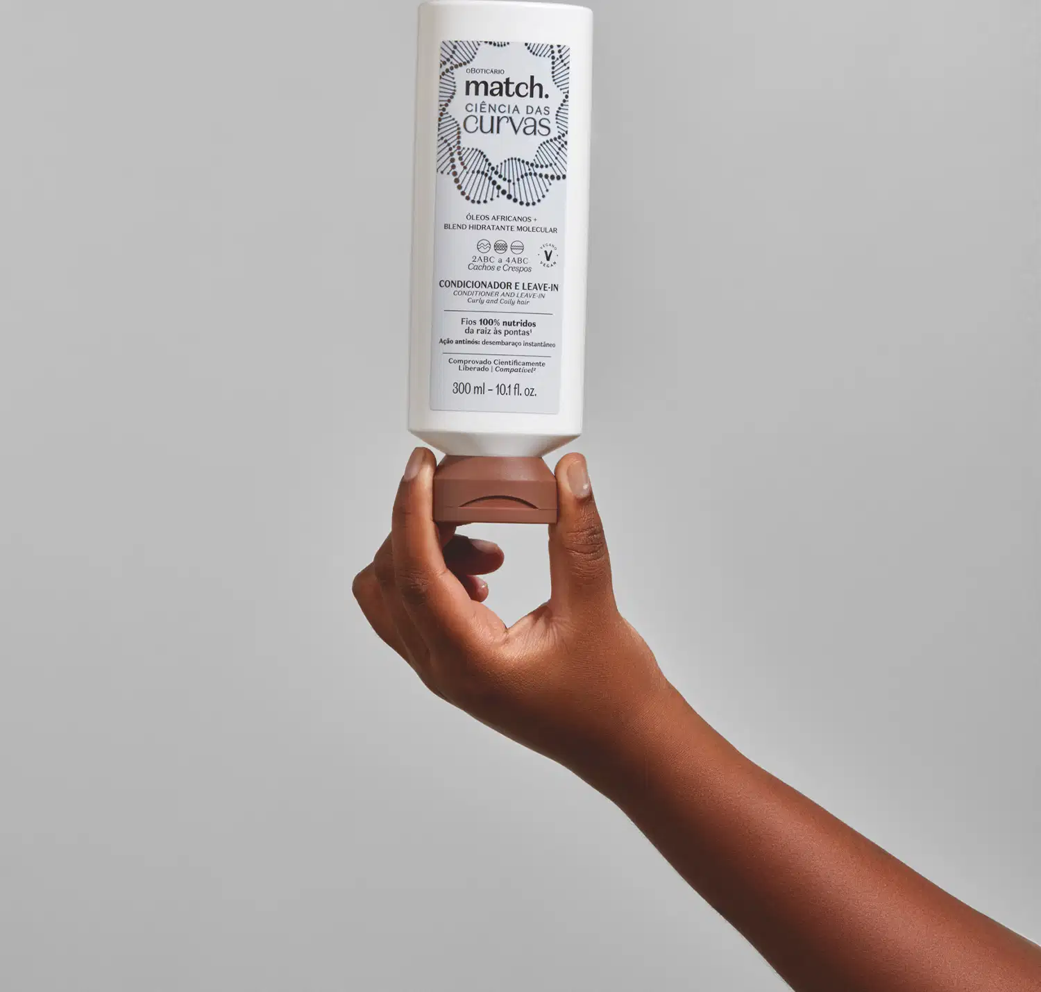 Imagem de uma embalagem branca de condicionador e leave-in Match Ciência das Curvas sendo segurada por uma mão em fundo cinza claro. O frasco tem a tampa marrom e informações impressas no rótulo em letras pretas.
