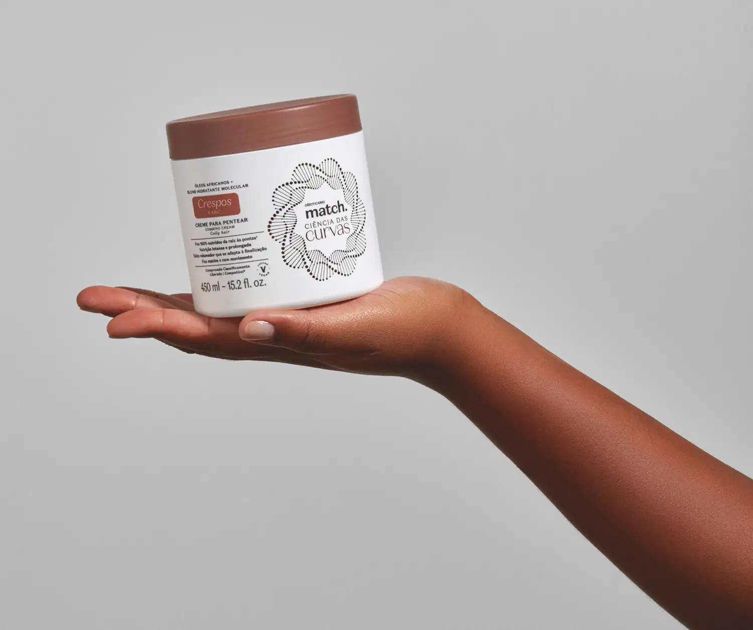 Imagem de uma mão segurando um pote branco com tampa marrom contendo creme para pentear da linha Match. Ciência das Curvas Crespos em fundo cinza.