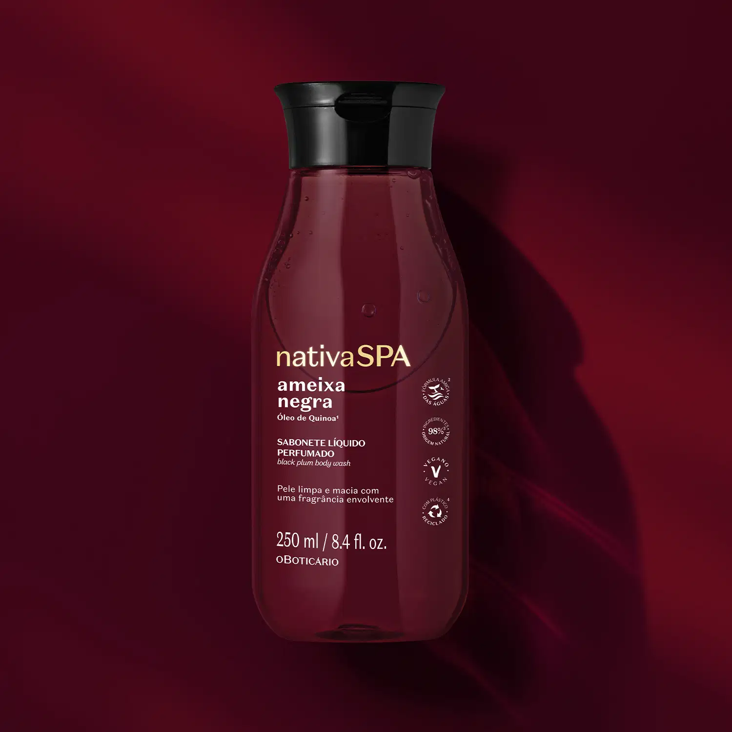A imagem mostra um frasco de sabonete líquido Nativa SPA Ameixa Negra, na cor vinho, com rótulo e informações em dourado. O produto possui tampa preta e está em fundo vermelho escuro, com sombras e reflexos que criam um ambiente elegante e sofisticado.