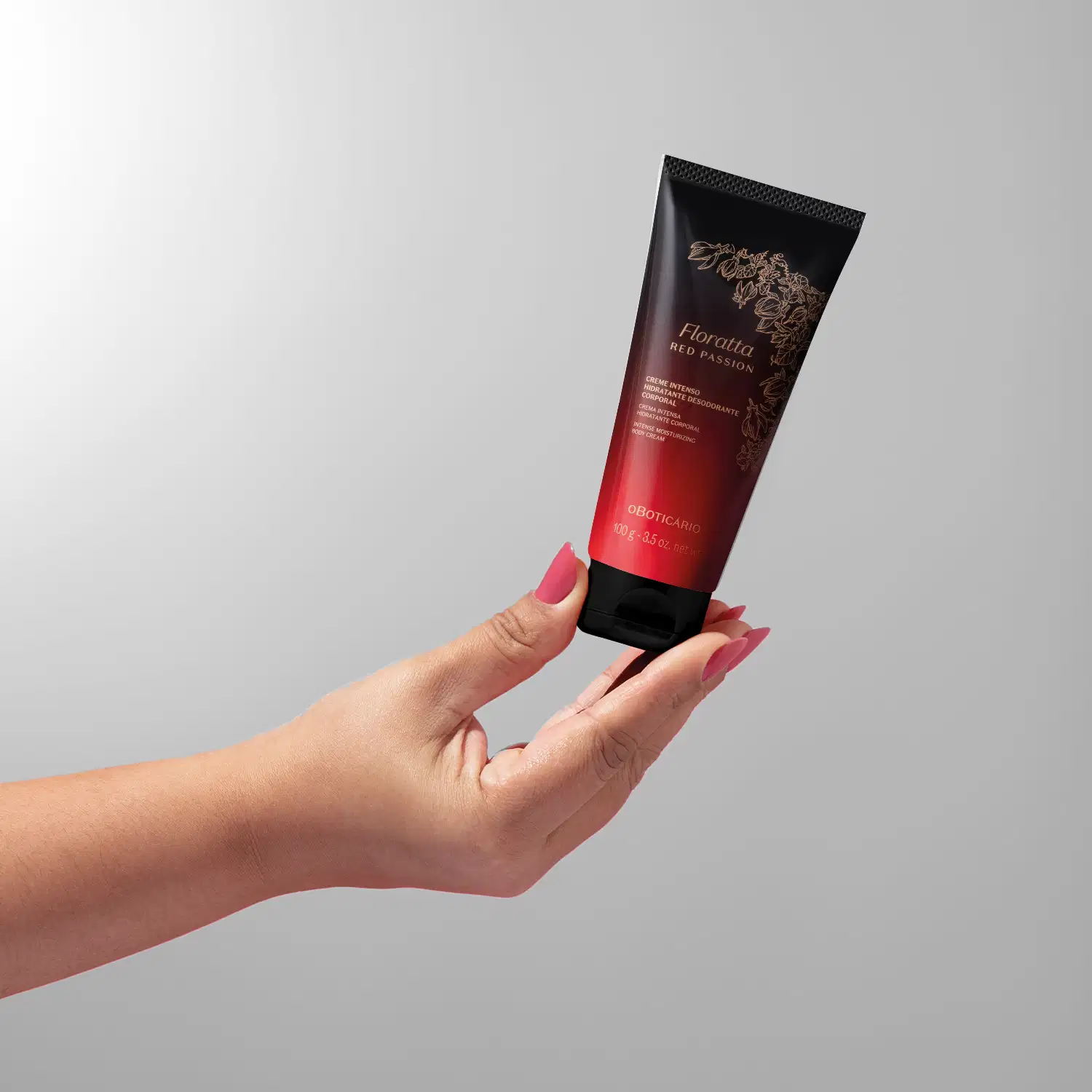 Imagem de uma mão feminina segurando uma bisnaga com degradê de preto e vermelho, tampa preta e rótulo dourado contendo creme intenso hidratante corporal da marca Floratta Red Passion. O fundo da imagem é em degradê de branco e cinza.