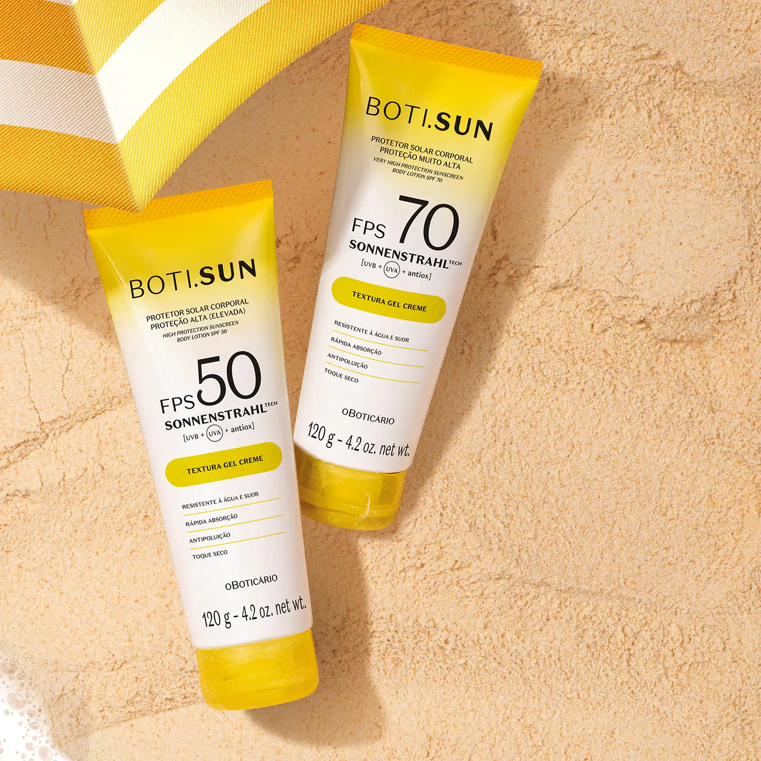 Dois tubos de protetor solar da marca Boti Sun, com FPS 50 e 70, sobre superfície bege simulando areia e parte de um guarda-sol amarelo e branco.