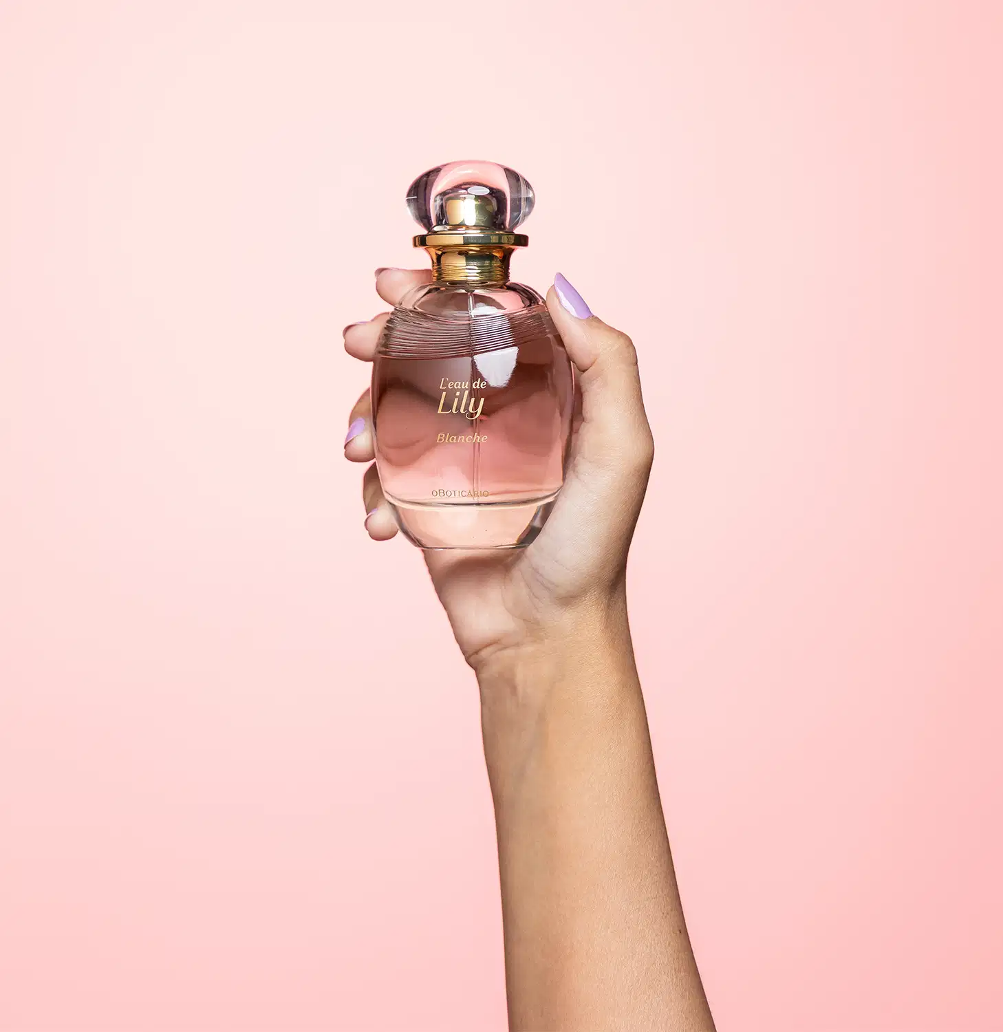 A cor predominante do frasco é um tom rosado, que transmite uma sensação delicada e feminina. A fragrância é segurada por uma mão feminina, com unhas pintadas em uma cor clara, indicando se tratar do desodorante colônia L'eau de Lily Blanche.
