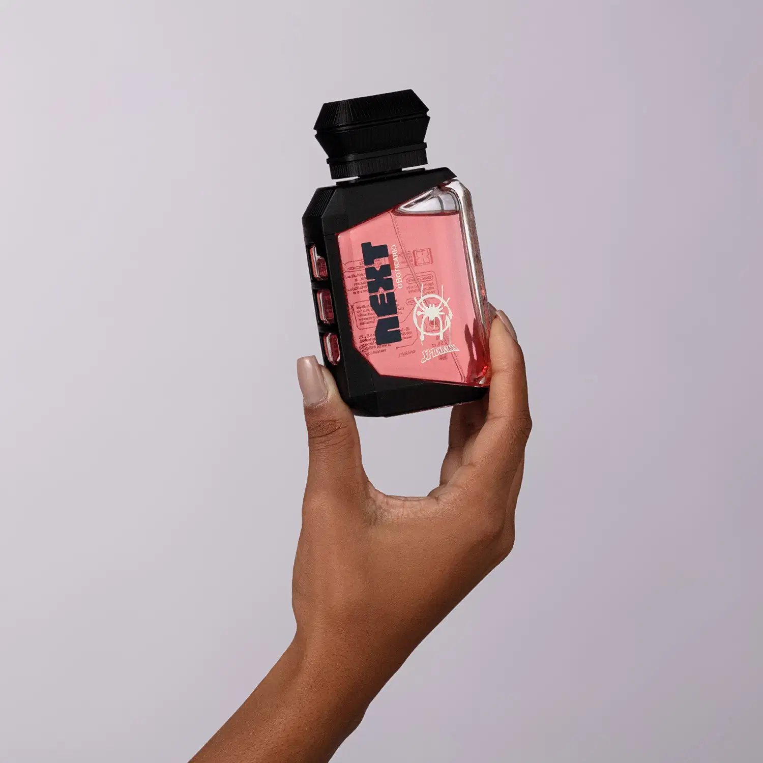 Frasco transparente de colônia infantil da marca Next, com detalhe geométrico preto e líquido rosa, exibindo logotipo da marca e da personagem Homem-Aranha sendo segurado por mão feminina negra contra fundo cinza.