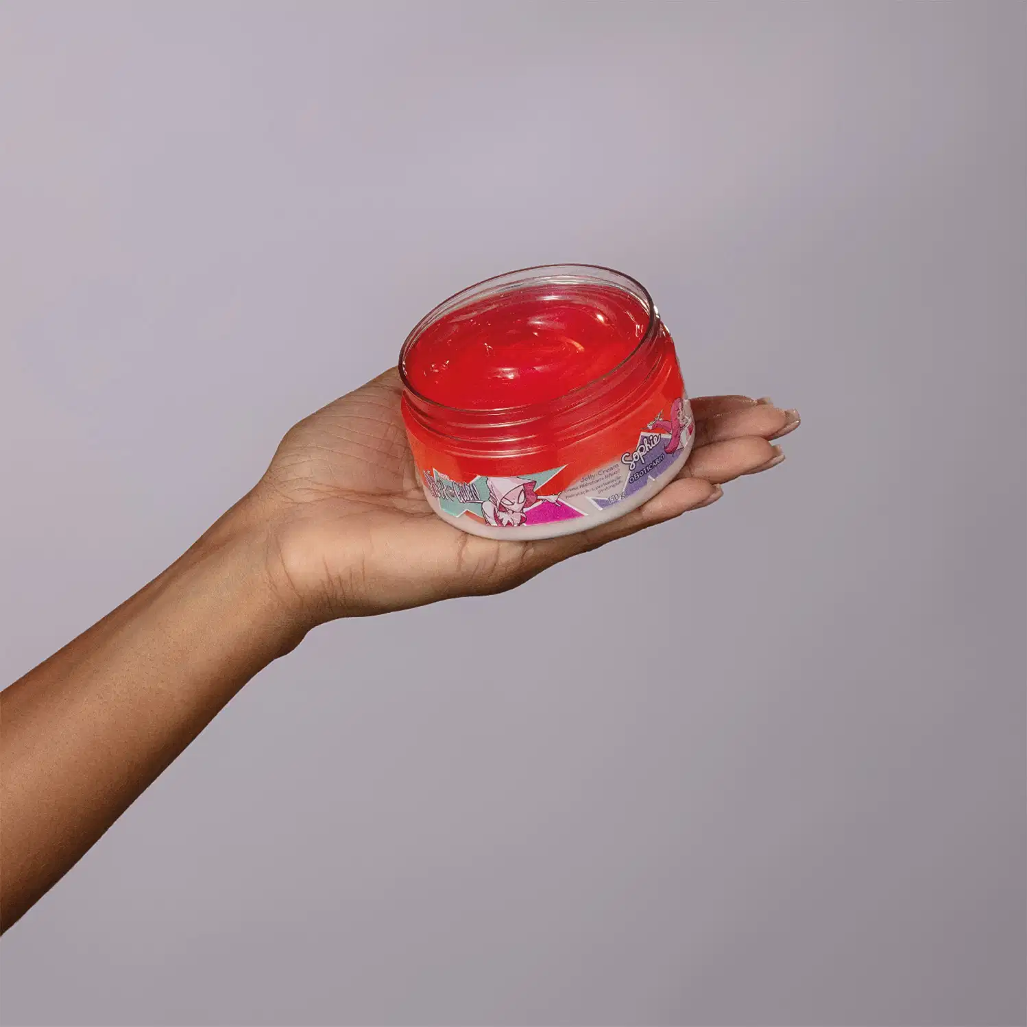 Pote aberto de creme hidratante da linha Sophie Marvel Spider-Gwen da marca O Boticário, com produto vermelho e gelatinoso dentro sobre mão estendida contra fundo cinza claro.