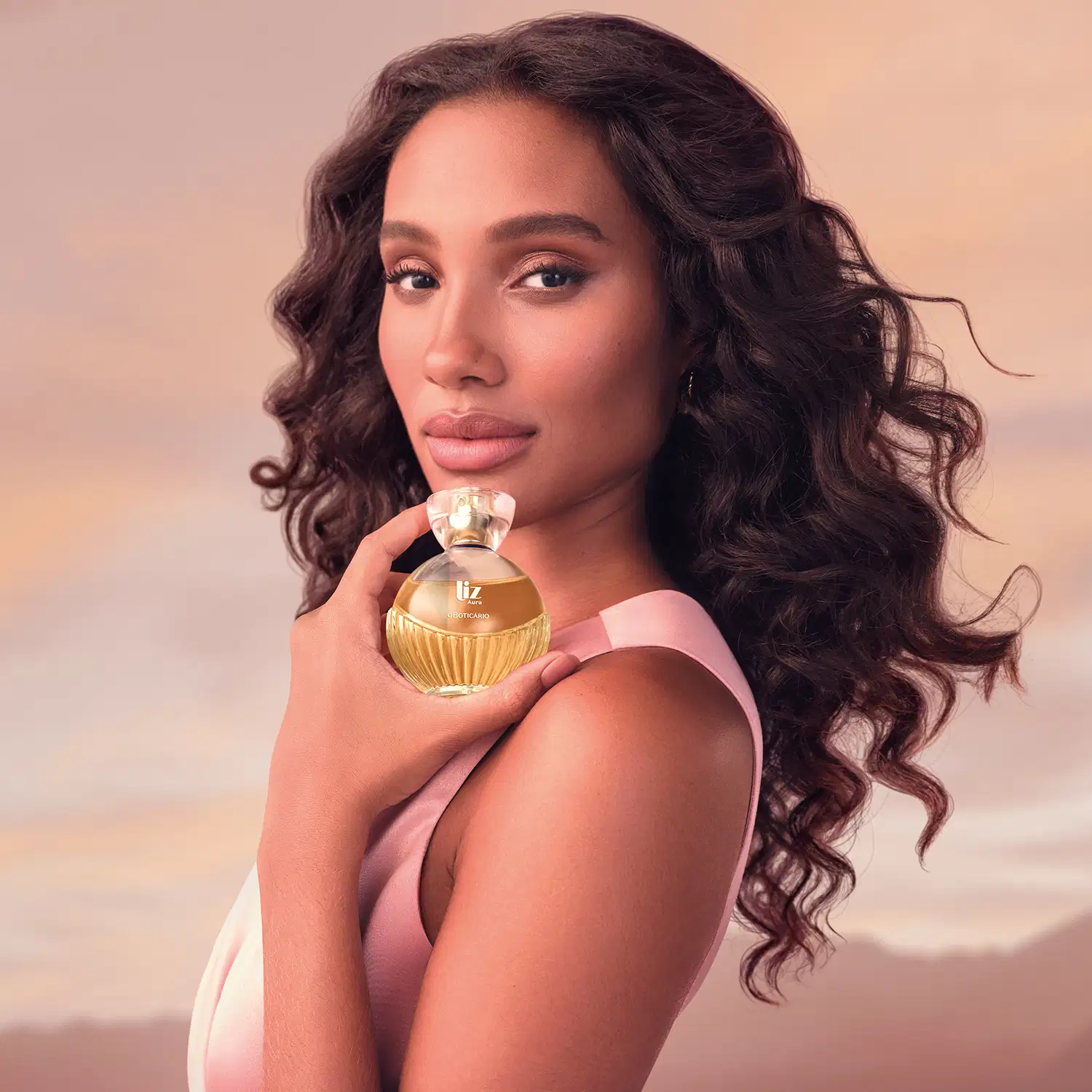 A mulher de cabelos cacheados e longos segura um frasco de desodorante colônia da marca Liz Aura em um ambiente com tons quentes de rosa e laranja, sugerindo um cenário de beleza e glamour.