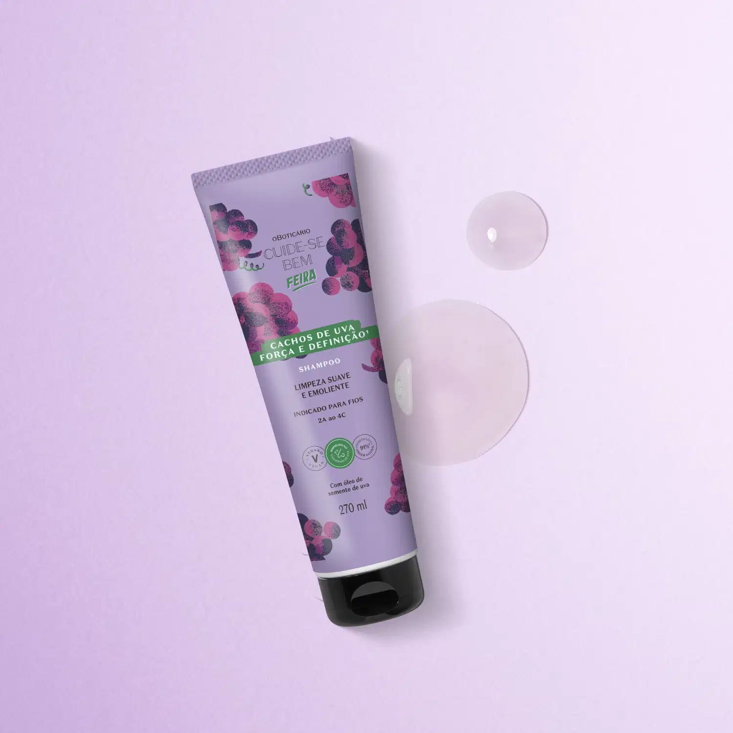 Imagem de um tubo de shampoo da marca Cuide-se bem Feira Cachos de Uva em embalagem roxa com ilustrações de uvas, sobre superfície lilás clara com duas gotas do produto, com textura gelatinosa e brilhante, ao lado.