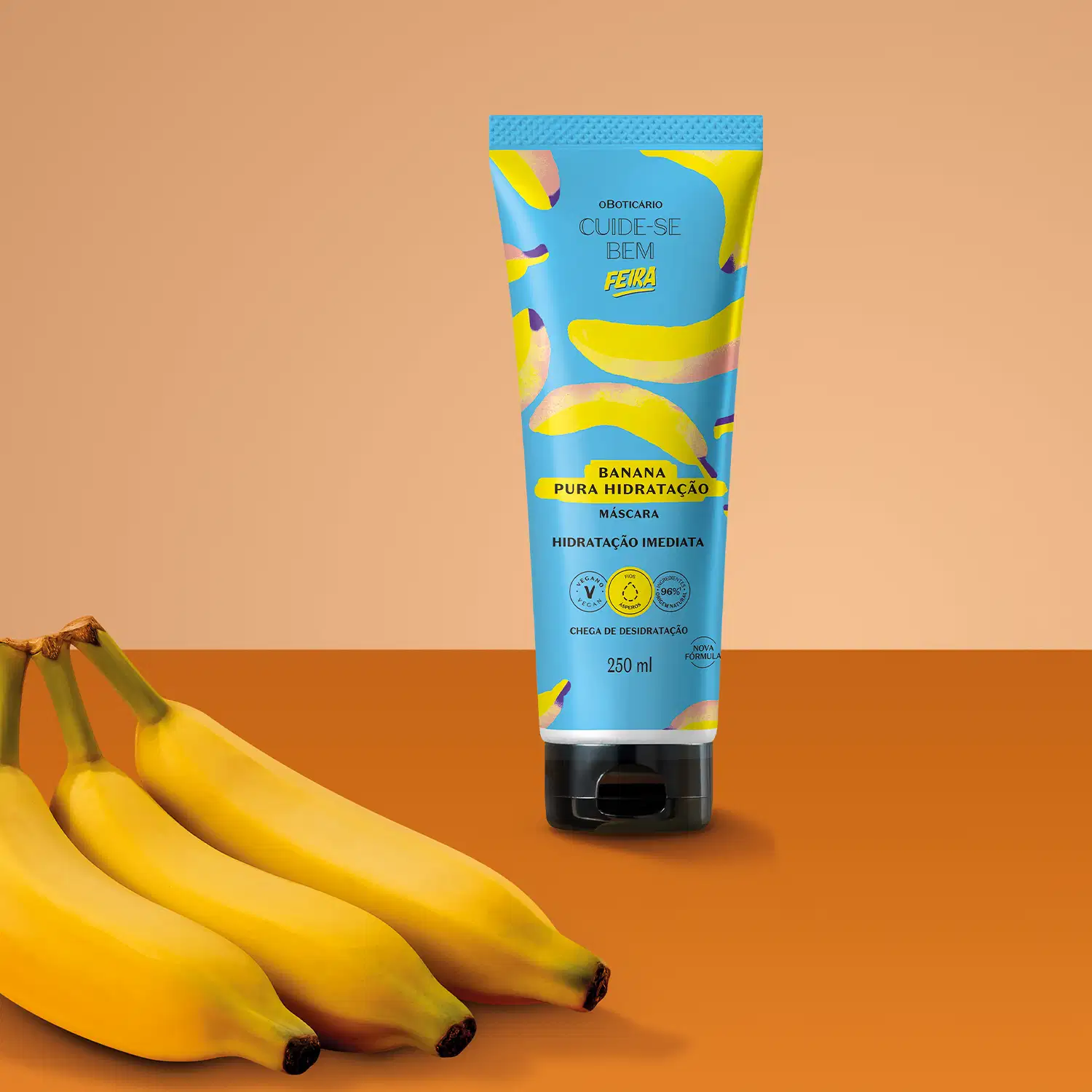 Imagem de um tubo azul com estampa de bananas amarelas de máscara capilar da marca Cuide-se bem Feira Banana, em fundo rosa e vermelho com bananas frescas ao lado.