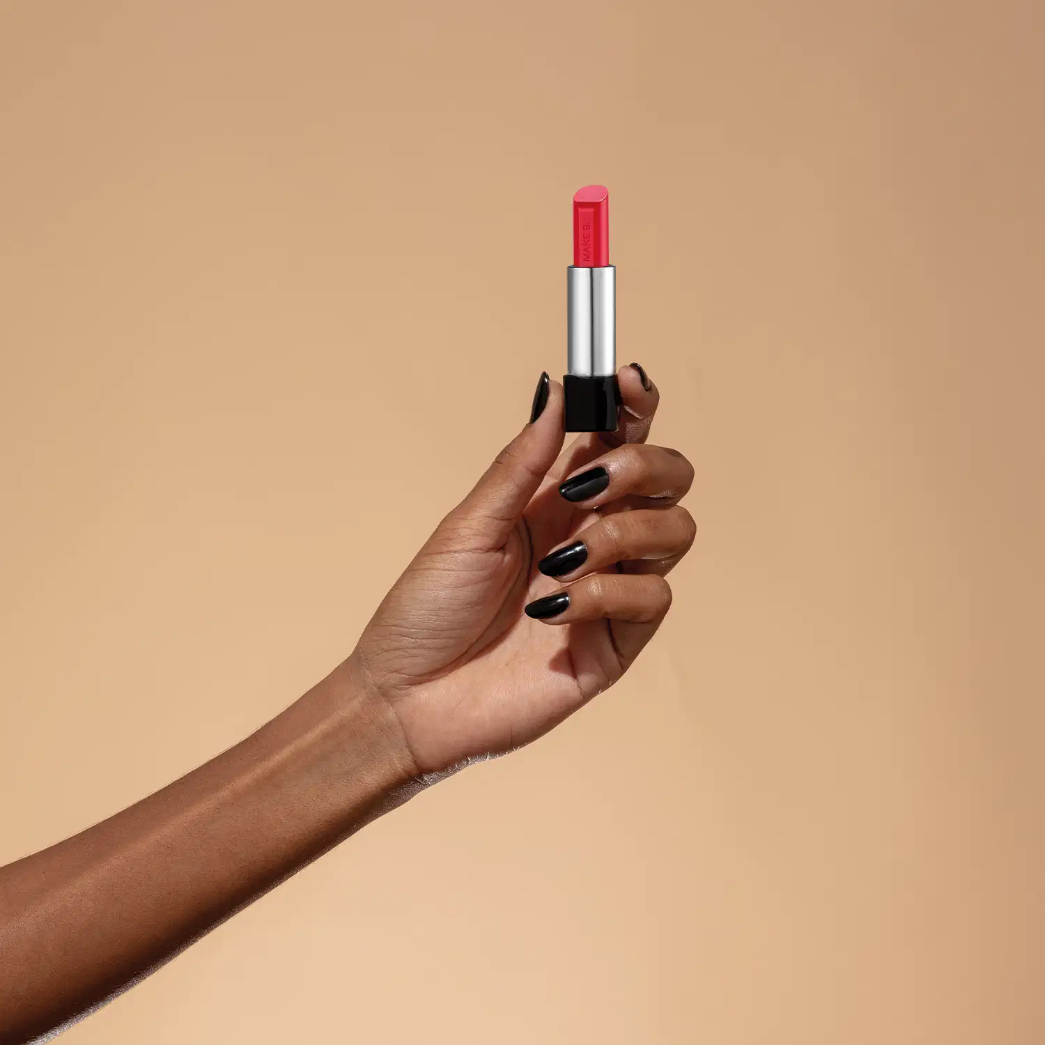 Uma mão feminina segura um balm labial vermelho da marca Make B. Lip Chronology Color Repair. O produto está centralizado contra um fundo laranja claro.