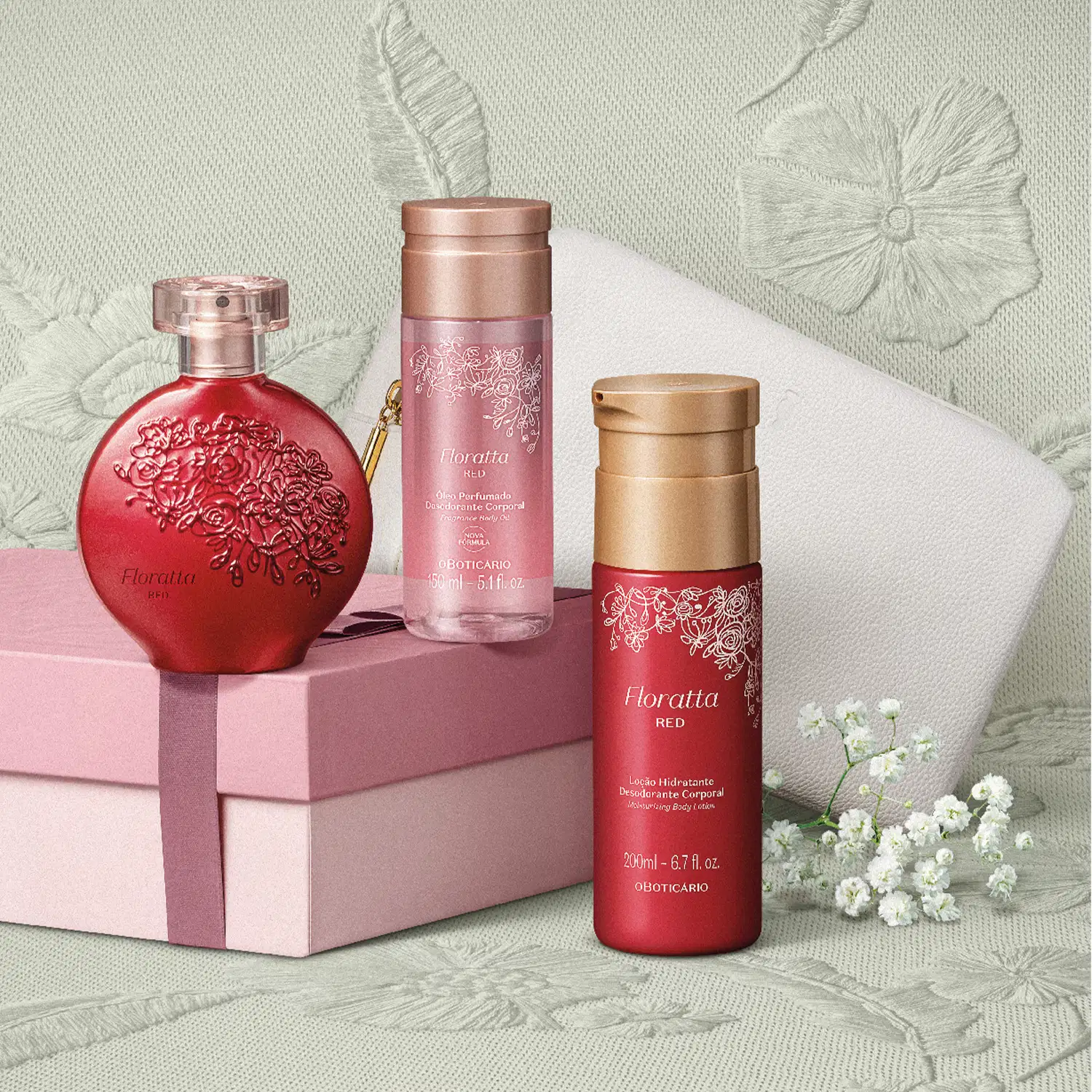Caixa rosa, nécessaire branco e três produtos Floratta Red, sendo um frasco de óleo perfumado corporal, vidro de desodorante colônia e embalagem de loção hidratante corporal, todos em cor vermelha com detalhes dourados em superfície bege com floral.