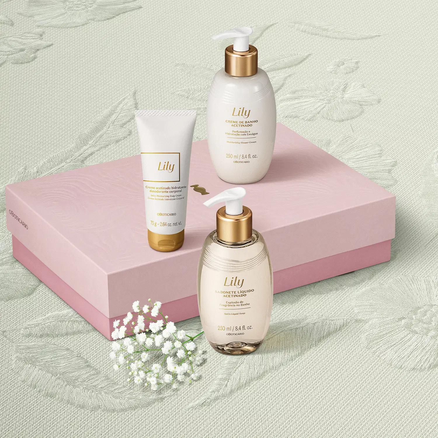 Caixa rosa com detalhe dourado e três produtos de cuidados corporais da marca Lily: bisnaga de creme acetinado corporal, frasco de creme de banho acetinado e outro frasco de sabonete líquido, todos em tons de dourado e branco em fundo bege com floral.