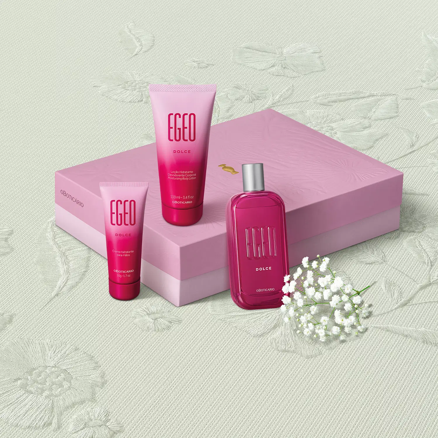 Caixa rosa com flores em relevo e três itens da marca Egeo Dolce, sendo uma bisnaga de creme hidratante para as mãos, outra de loção hidratante corporal e um vidro com tampa prateada contendo desodorante colônia com flores brancas ao redor em fundo bege.