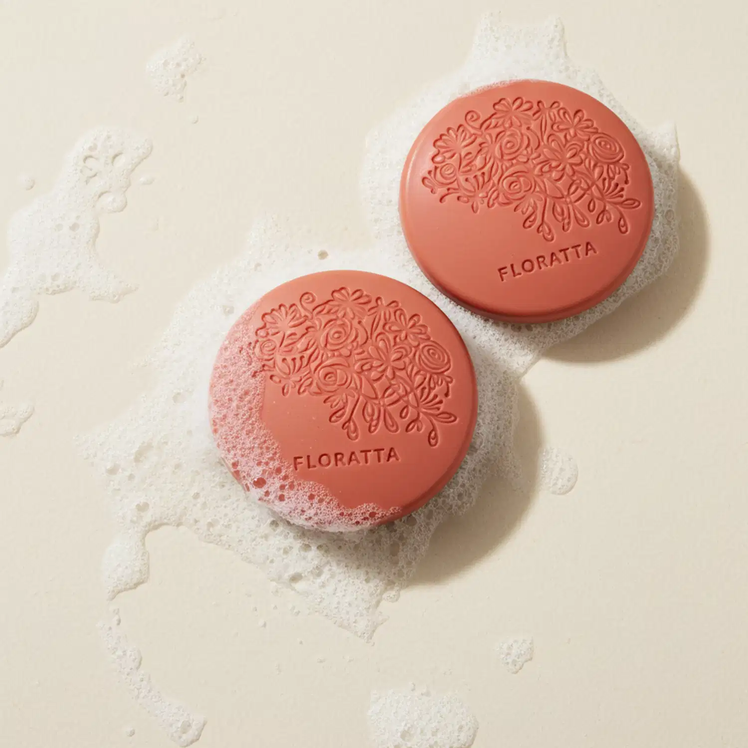 IImagem de dois sabonetes  redondos na cor rosa da marca Floratta Red em superfície branca molhada com espuma ao redor dos produtos.