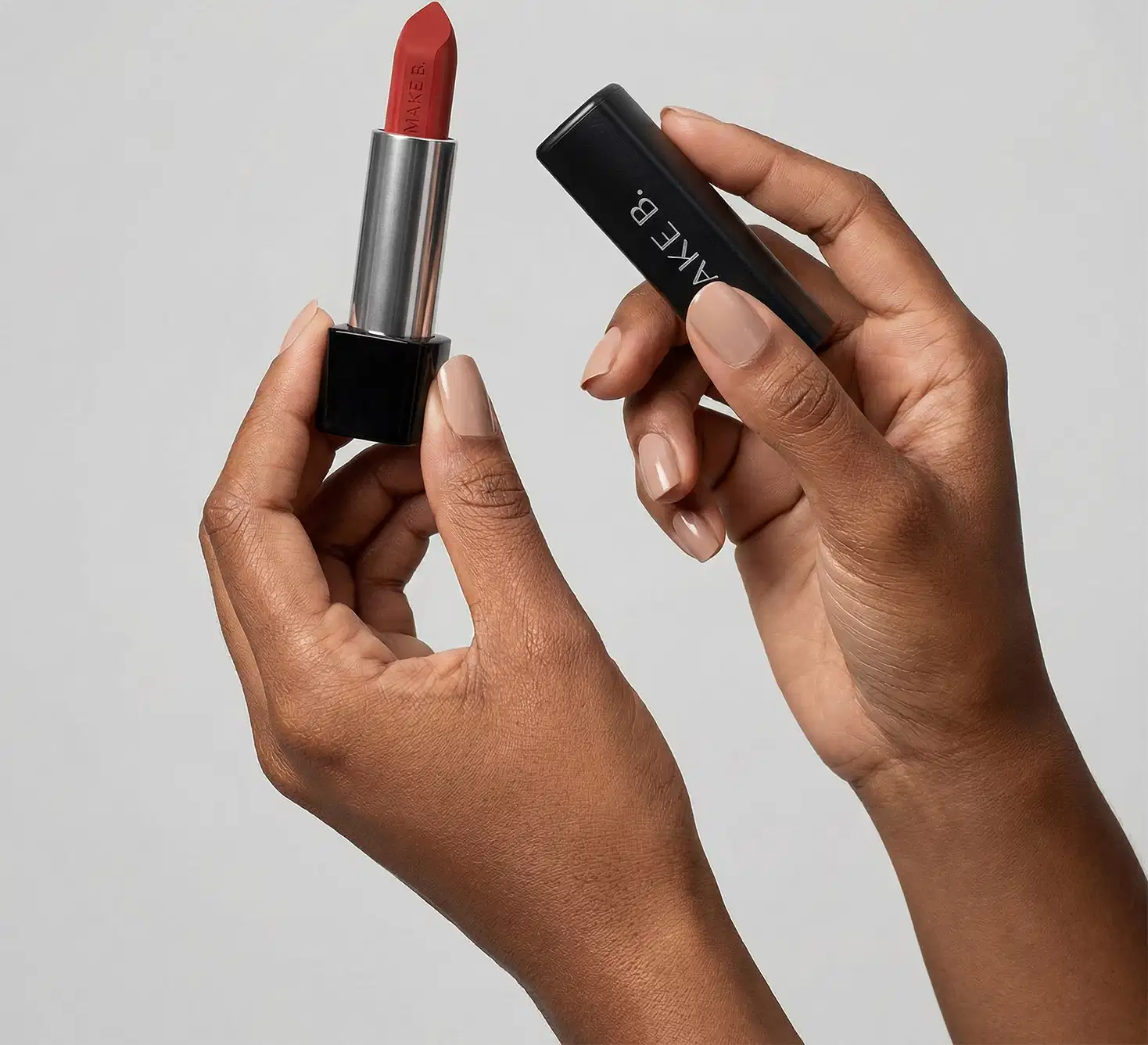 Mãos femininas segurando a tampa preta de um batom da marca Make B. na mão direita e a base do batom com a bala aparente na mão esquerda, em um ambiente minimalista e claro.