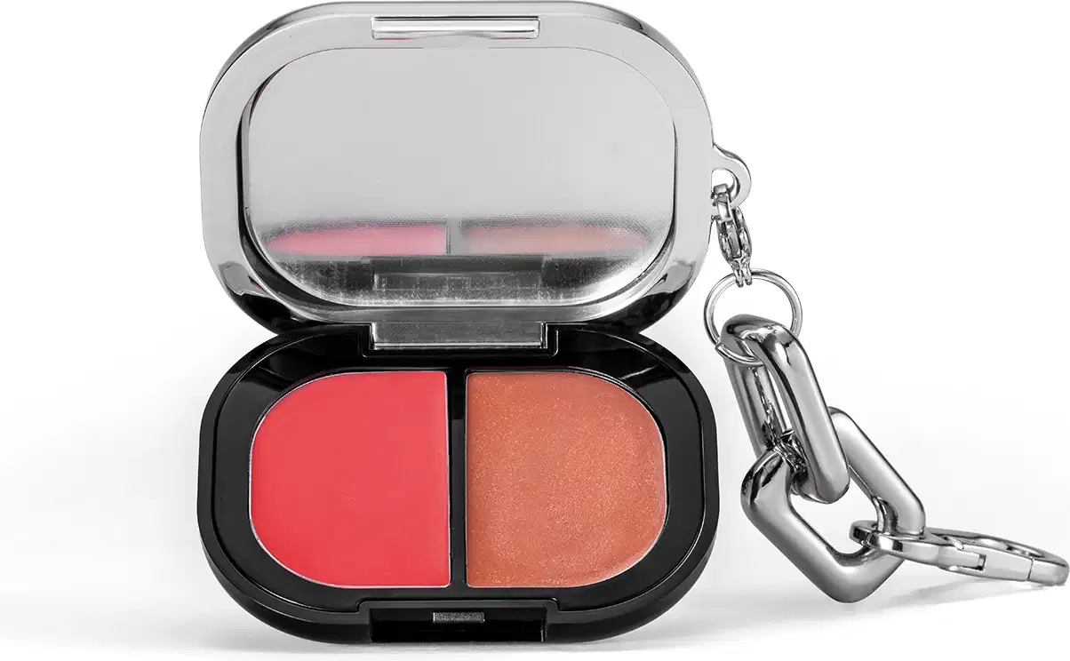 A imagem mostra um estojo de blush compacto com duas cores, uma rosa e uma bege, em embalagem preta e prateada com um gancho metálico.