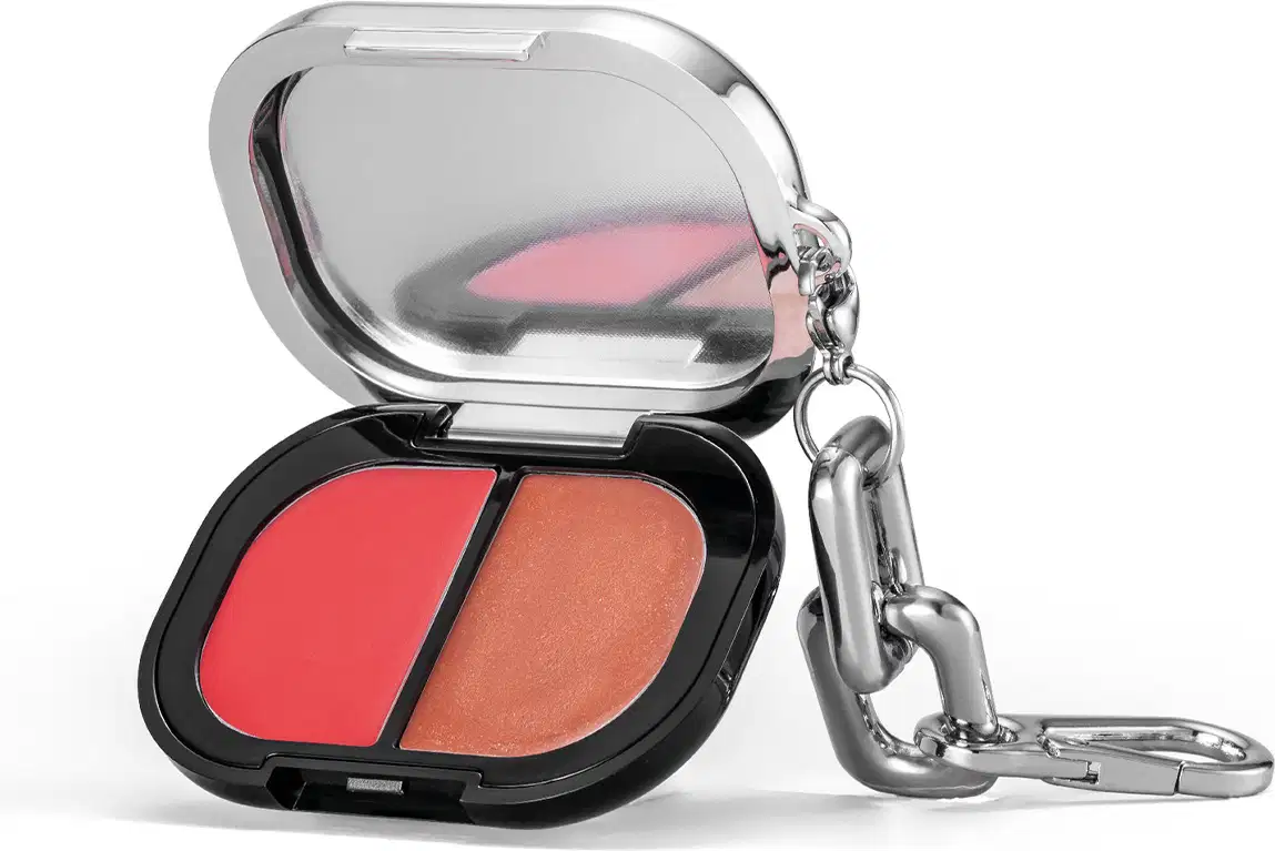 A imagem mostra o estojo do blush compacto aberta, apresentando as duas cores do produto em uma embalagem preta e prata. Ele possui um pingente metálico em formato de gancho preso à tampa.