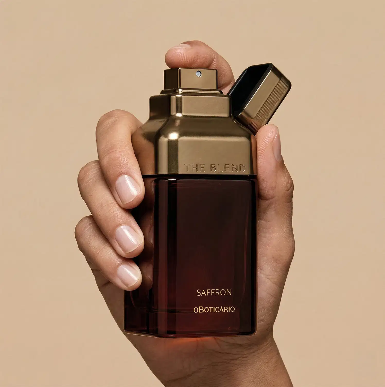 Imagem de uma mão segurando um frasco de fragrância da marca The Blend, com a fragrância Saffron, em um ambiente neutro de fundo bege, destacando o design elegante e sofisticado do produto.