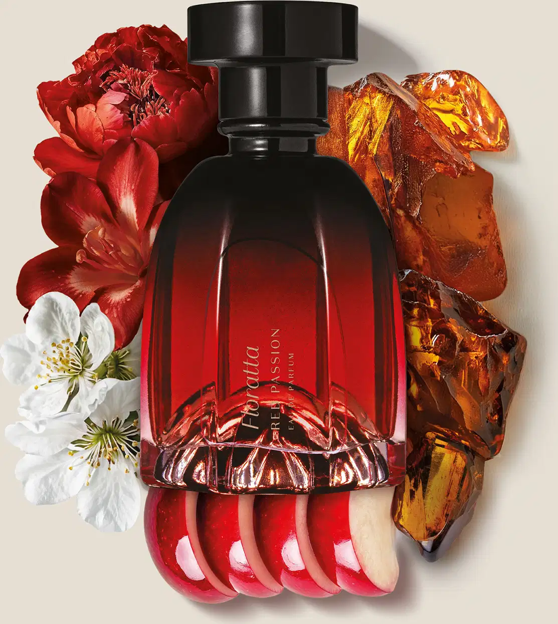 Frasco de item de perfumaria com formato de uma taça invertida feito em vidro, com predominância de cores preta e vermelha cercado por flores vermelhas e brancas, pepitas de âmbar e fatias de maçã vermelha sobre superfície branca.