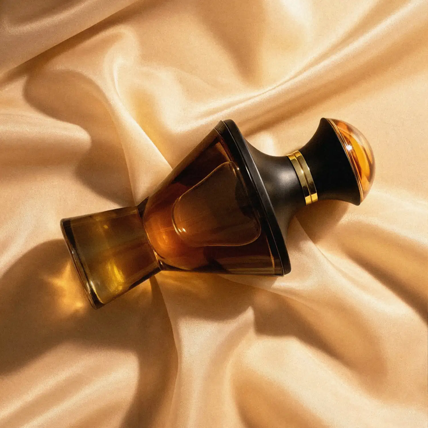 Imagem de um frasco de Eau de Parfum da marca Hadiya  em vidro âmbar com detalhes dourados sobre tecido bege com brilho suave, em ambiente iluminado e aconchegante.