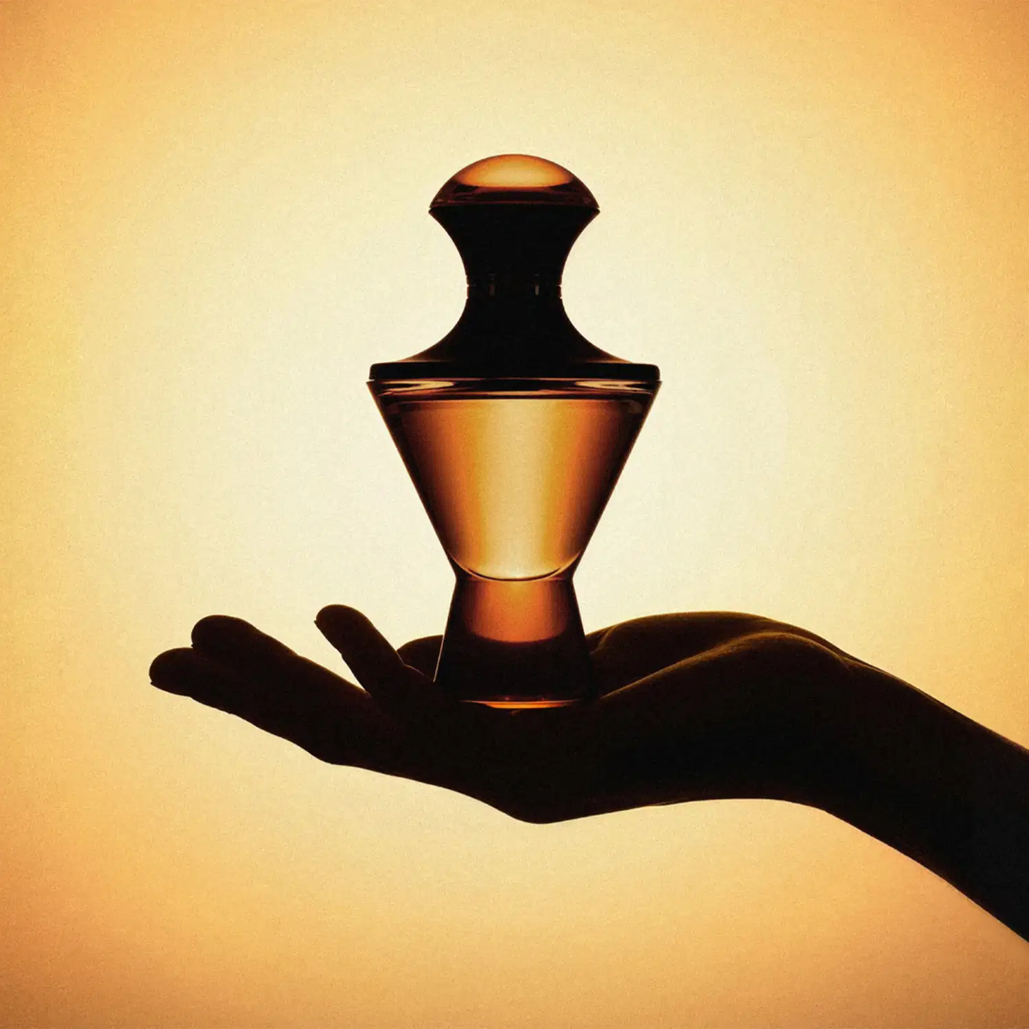 Imagem de uma mão segurando um vidro elegante e sofisticado de Eau de Parfum da marca Hadiya em fundo marrom iluminado. Seu formato transmite luxo e exclusividade. mostrando se tratar de uma fragrância cheia de personalidade.