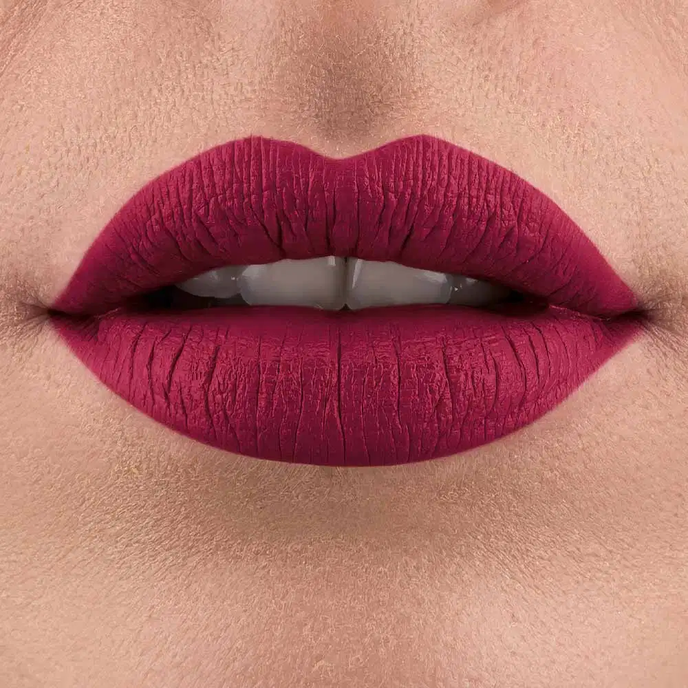Lábios entreabertos pintados com batom rosa escuro em close-up.  Vê-se um pequeno pedaço de um rosto feminino de pele branca, com destaque para os lábios.
