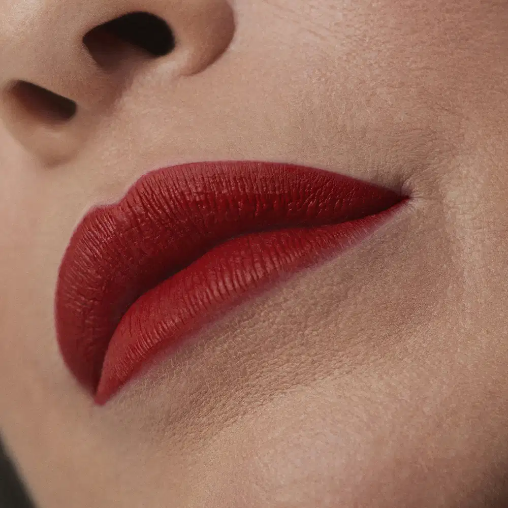 Imagem de um rosto feminino em close-up, evidenciando os lábios com o batom líquido Make B. em tom vermelho bem forte. A boca está fechada com um semblante sério.