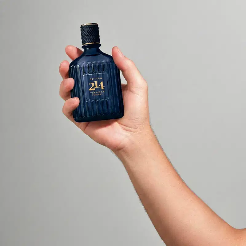 Imagem de um braço com uma mão segurando uma embalagem da fragrância da marca Botica 214, em um fundo cinza claro. A embalagem possui um design elegante com detalhes em preto e dourado, sugerindo um produto de luxo e sofisticação.