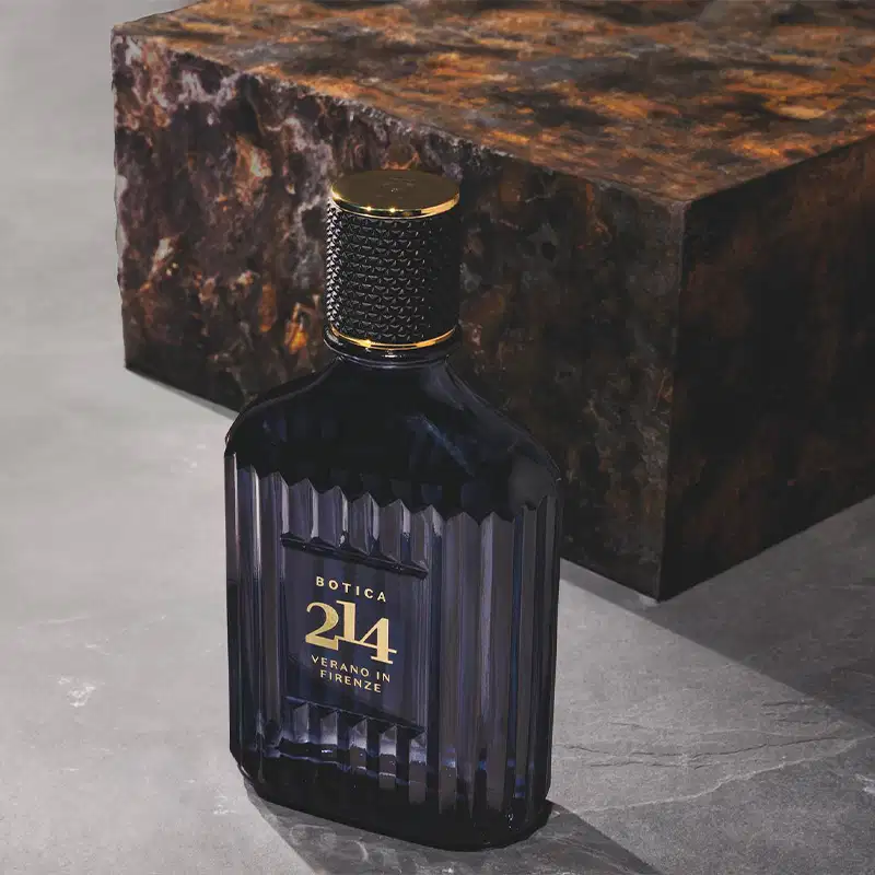 Imagem de um ambiente elegante e sofisticado, com uma garrafa de fragrância da marca Botica 214, em embalagem escura e com detalhes dourados, destacada em um fundo cinza. Atrás da embalagem há uma caixa em tons quentes, sugerindo um ambiente de luxo.
