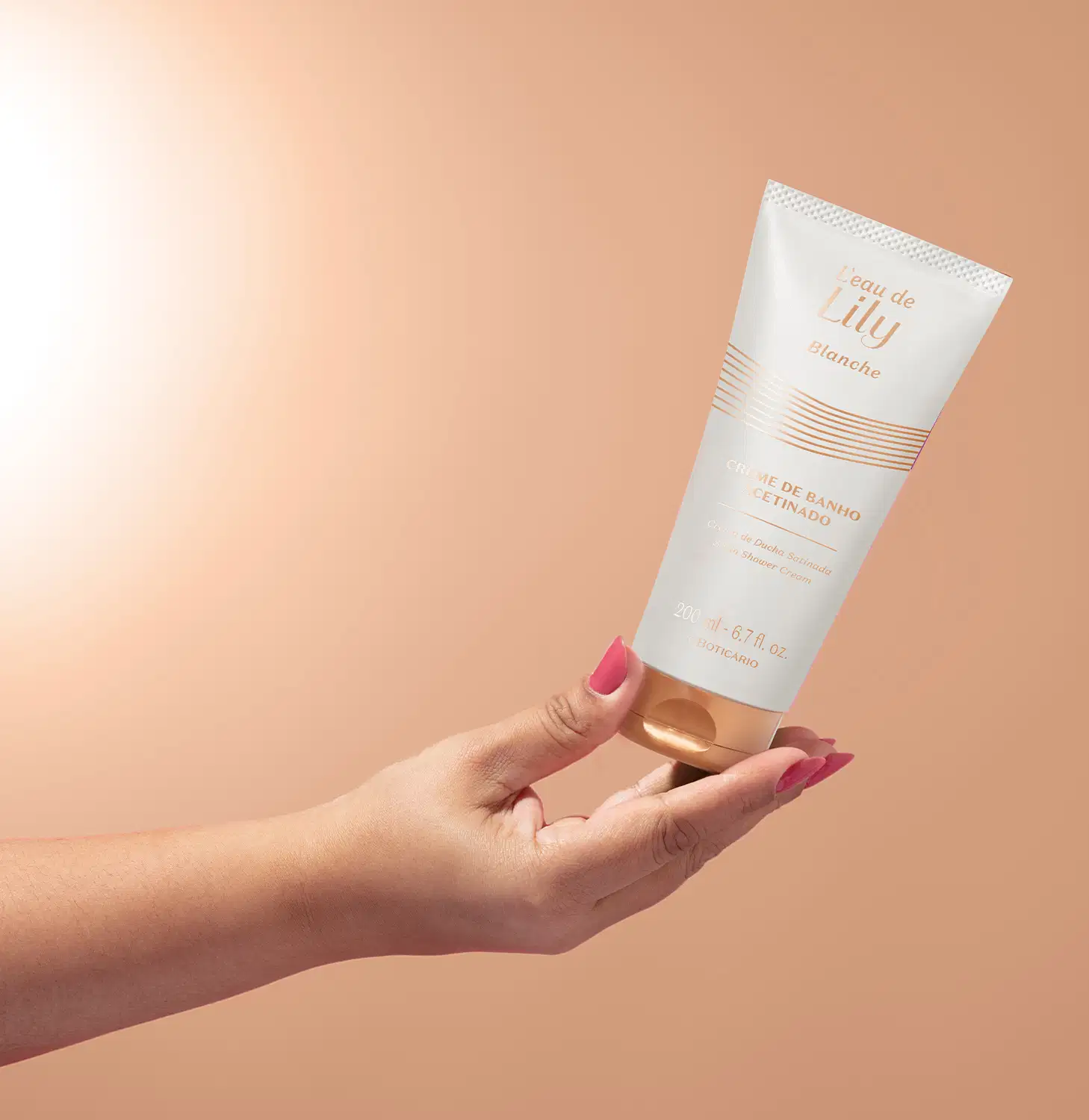 Imagem de uma mão segurando um frasco branco com tampa e rótulo dourados de creme de banho acetinado da linha L'eau de Lily Blanche em fundo com degradê de branco e rosa.