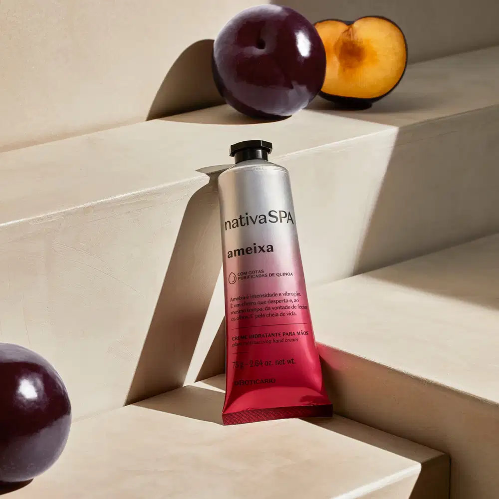 Imagem da embalagem prata e rosa do creme hidratante para as mãos Nativa SPA Ameixa sobre superfície clara, com ameixas roxas inteiras e cortadas ao meio ao redor, em ambiente minimalista e neutro.