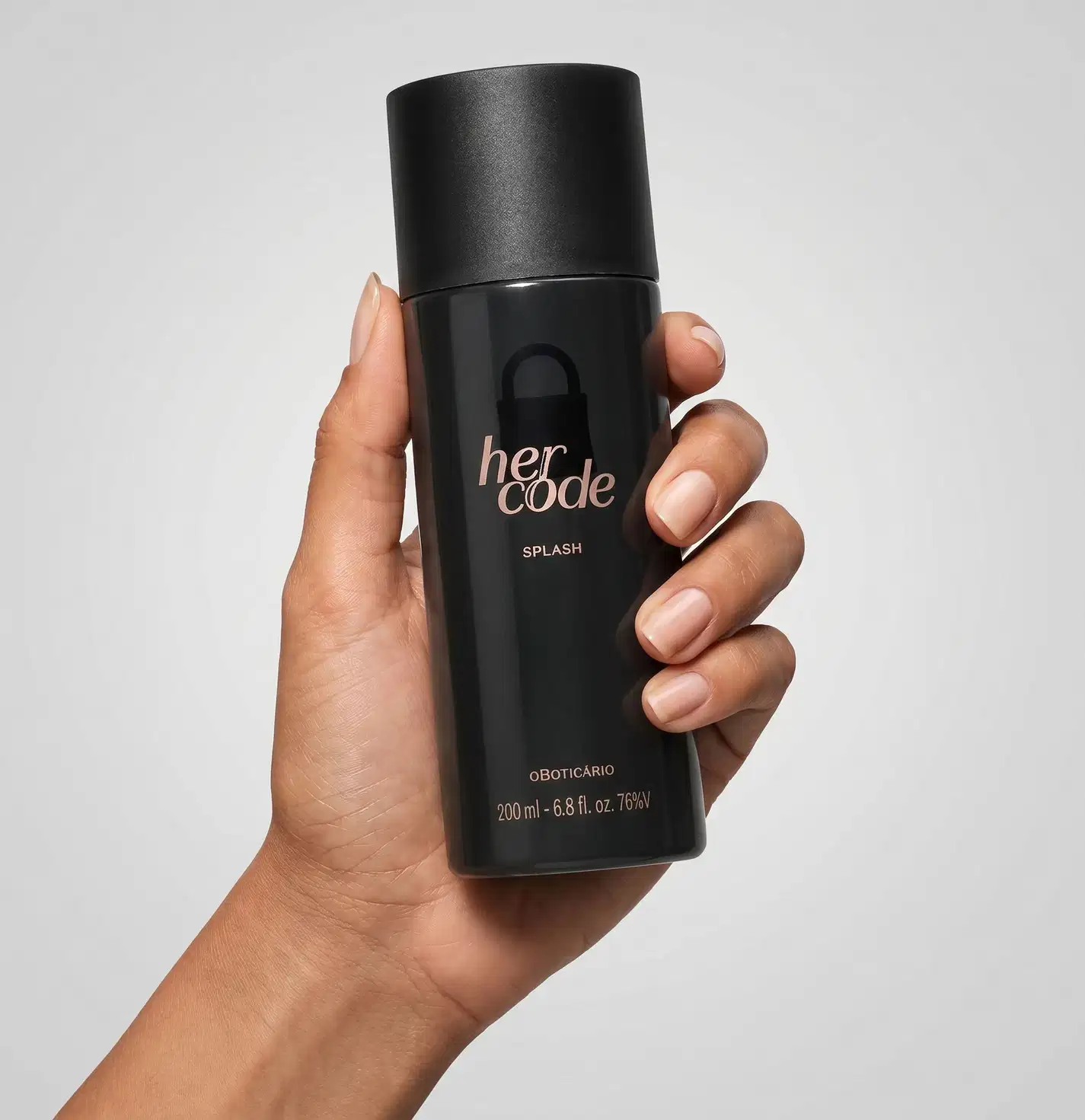 Frasco preto de item de perfumaria da marca Her Code, com logotipo  de uma discreta silhueta de cadeado e nome da marca, sendo segurado por mão feminina de pele branca sobre fundo branco.
