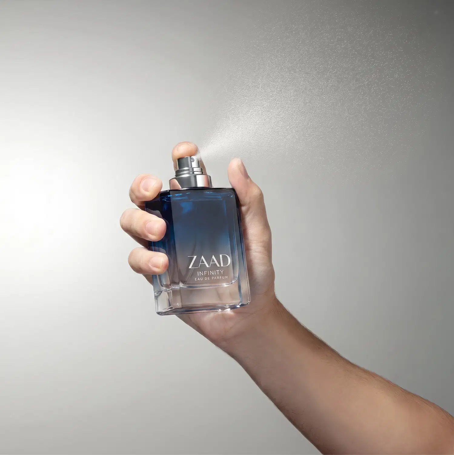 Frasco de item de perfumaria da marca Zaad Infinity em mãos contra fundo branco, com névoa de pulverização saindo do borrifador.